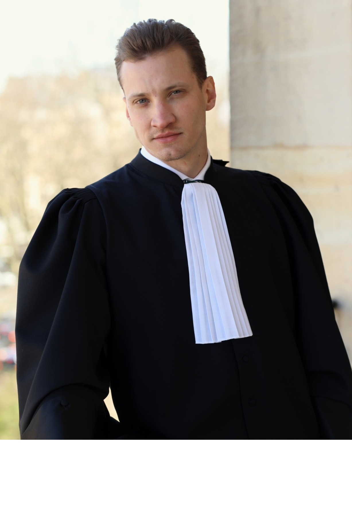 Jeune homme portant une robe d'avocat, debout devant un bâtiment en pierre, avec un fond flou d'arbres, regardant l'objectif.