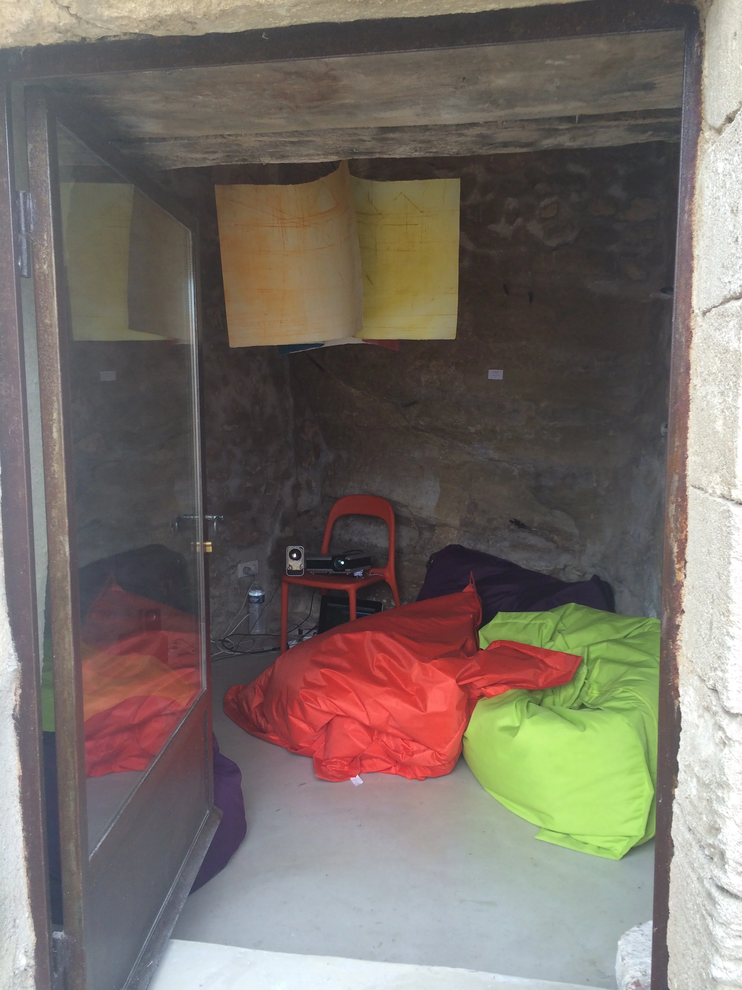 Interior of a small rock shelter with colorful sleeping bags on the ground, a red chair, a small table with a camera, and a map hanging on the wall.