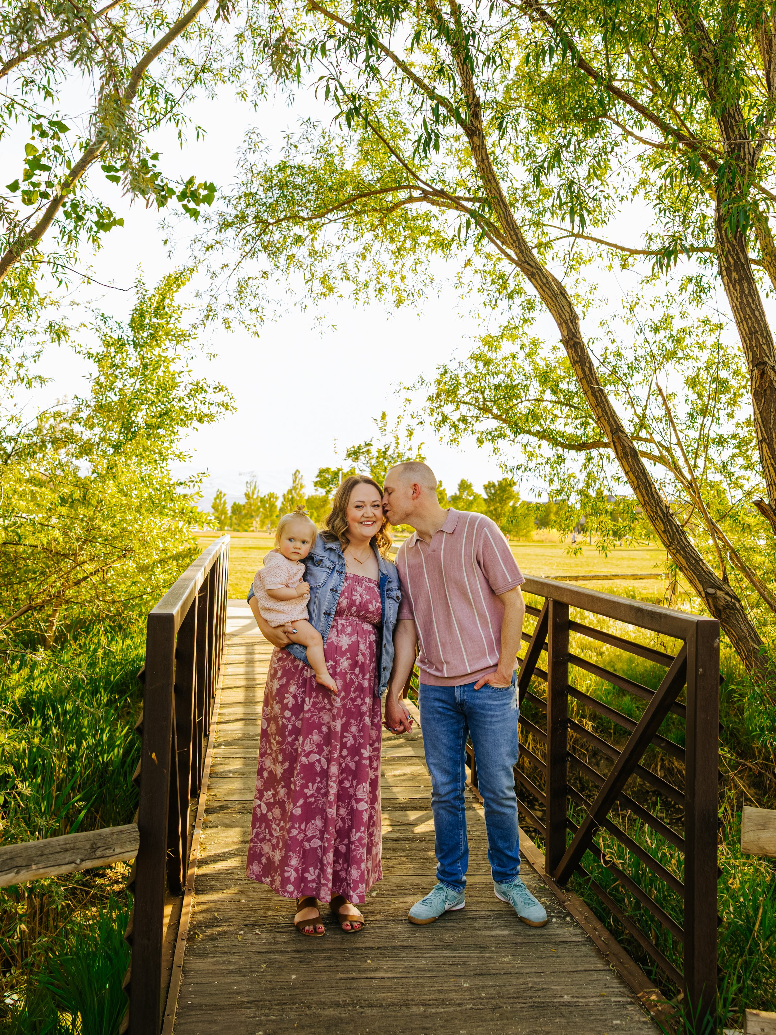 A Family of three smiling and posing on a wooden bridge in a park during golden hour.