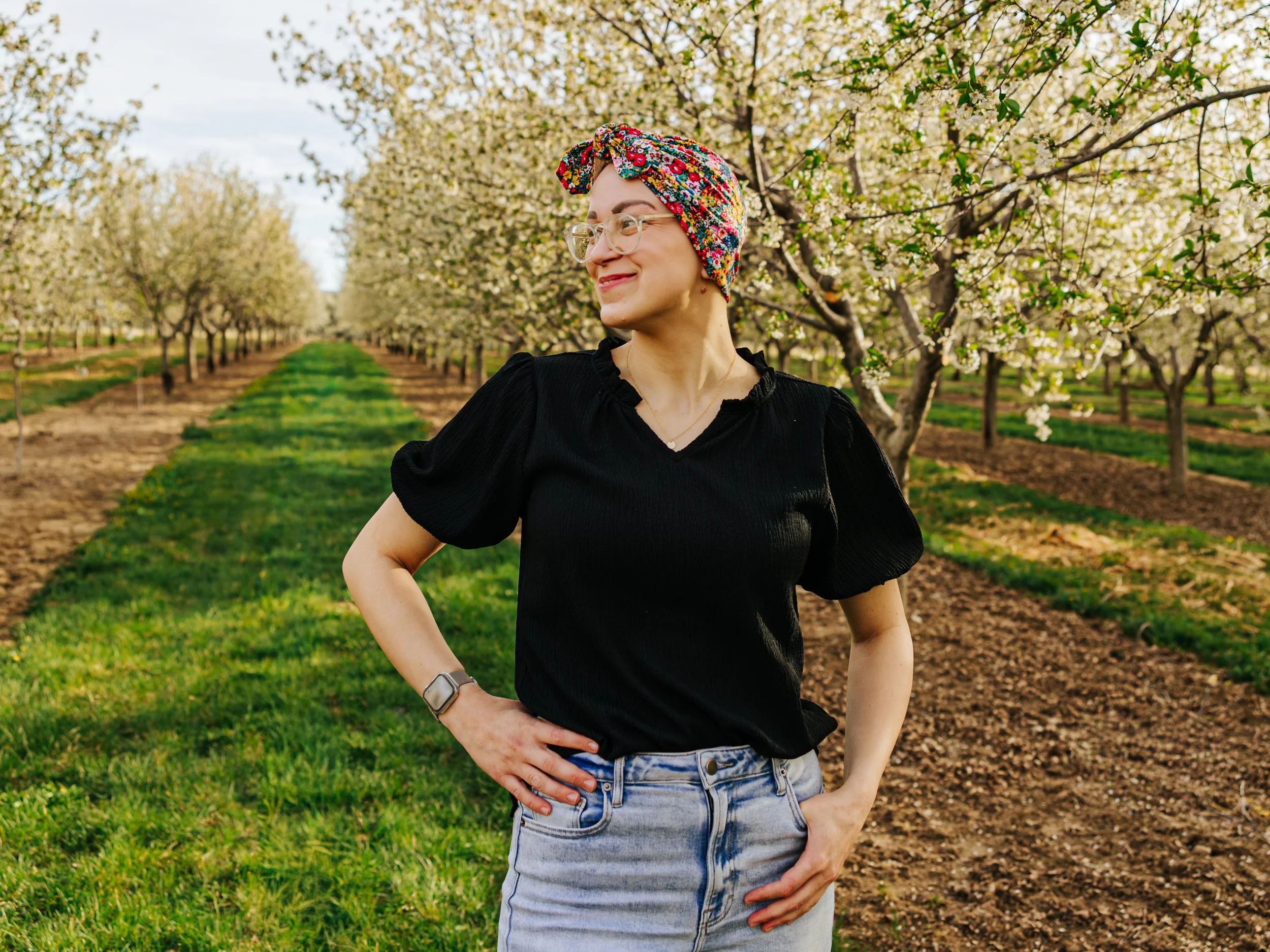 A woman smiling in a black top, light blue jeans, and a colorful floral headscarf standing in an orchard with white blooming trees and green grass.