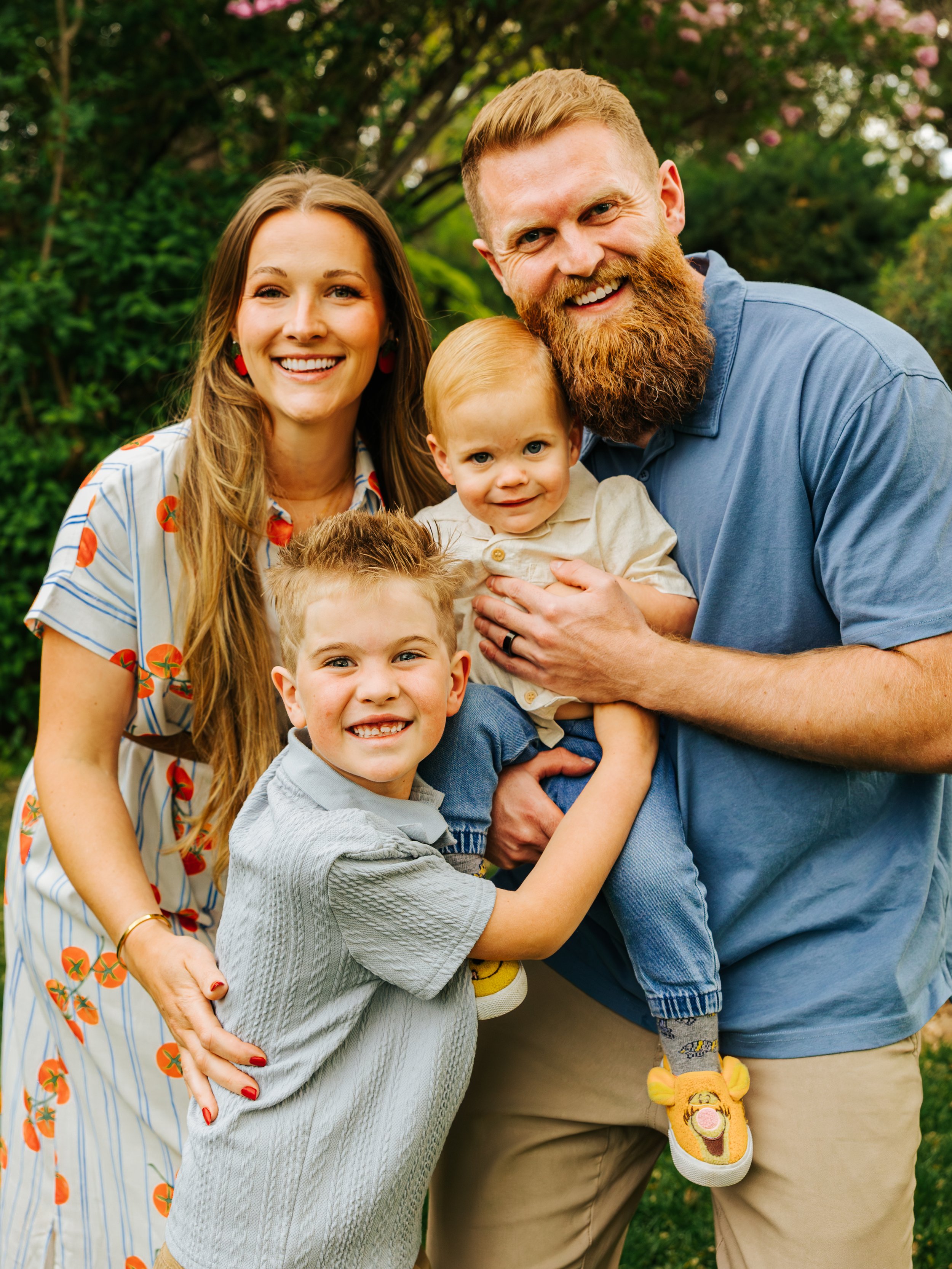 A family of five outdoor, smiling, standing close together, with lush green trees and pink flowers in background.