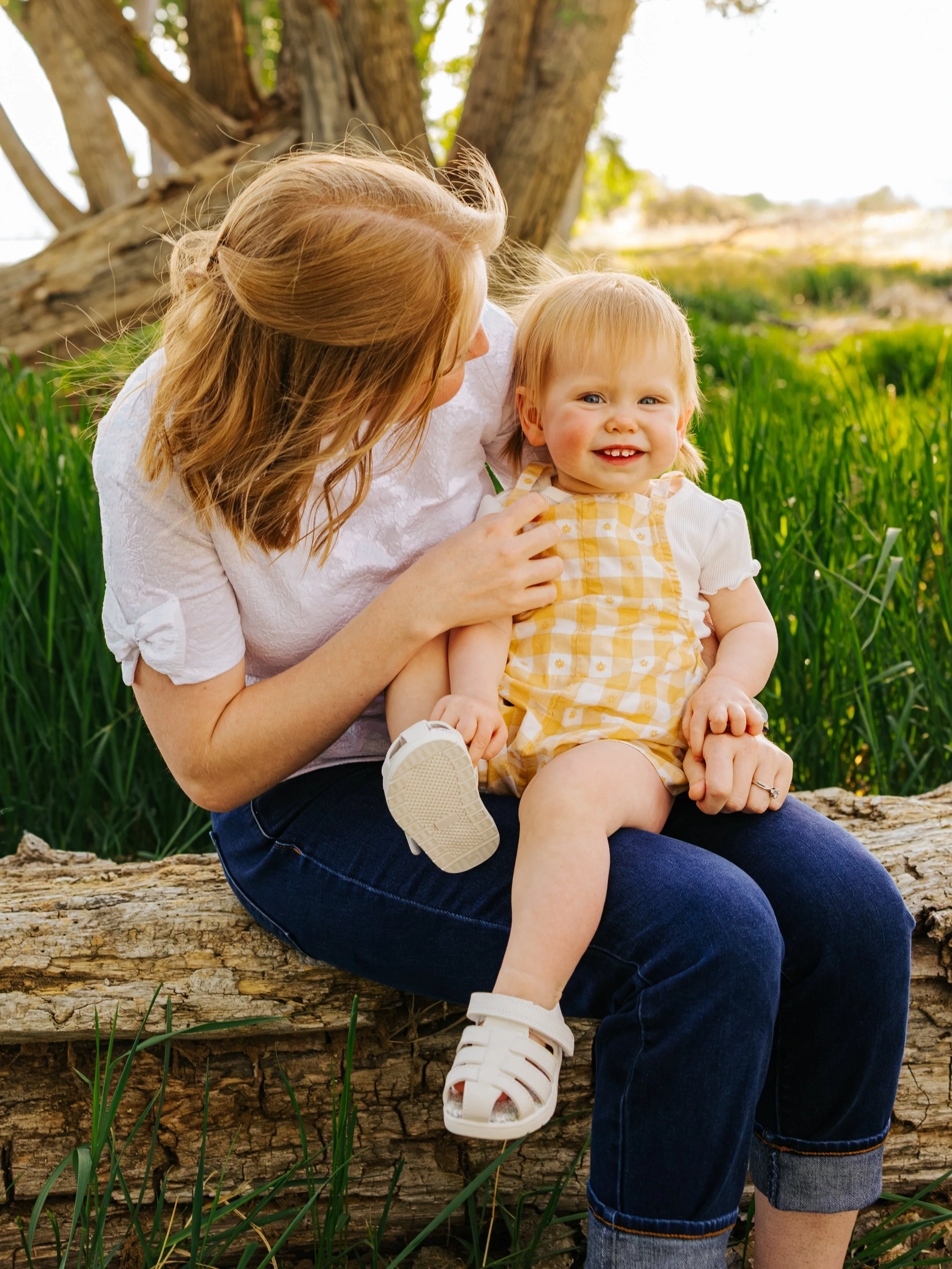 A woman with long reddish hair and a young girl with blonde hair sitting on a decayed tree trunk outdoors, with green grass, trees, and a water body in the background. The woman is looking at the girl, who is smiling at the camera.