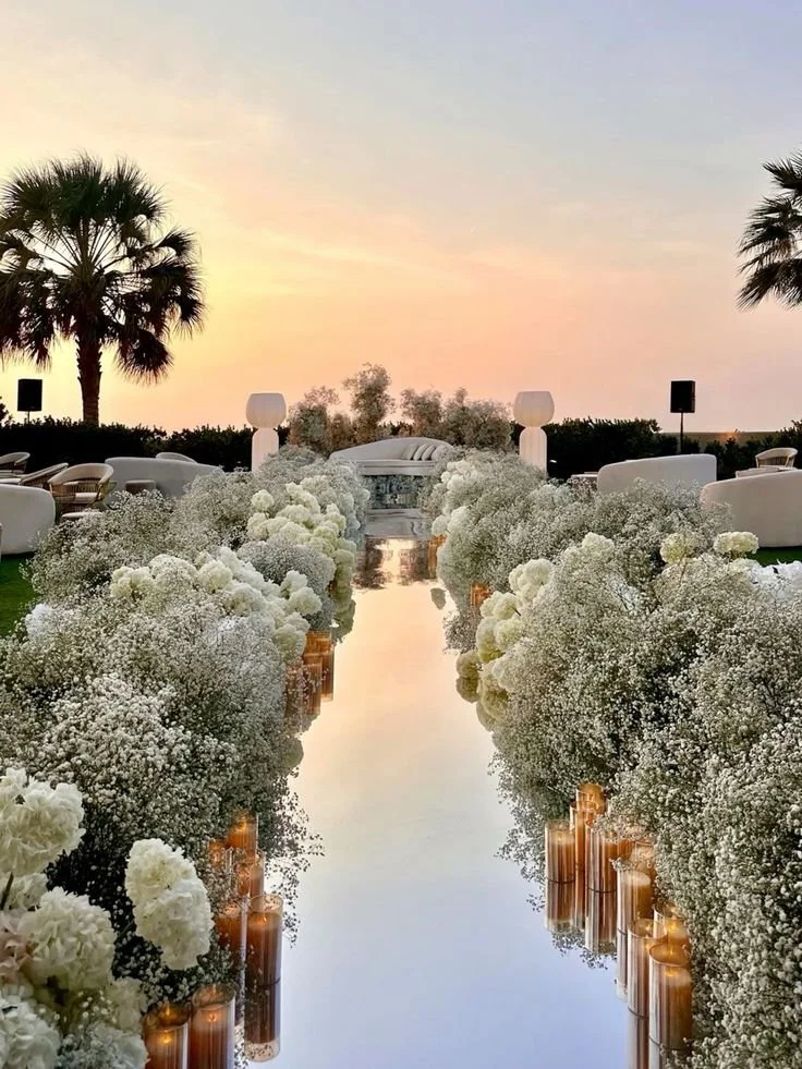 A floral aisle with white flowers and candles leads to an outdoor wedding altar under a pastel sky with palm trees.