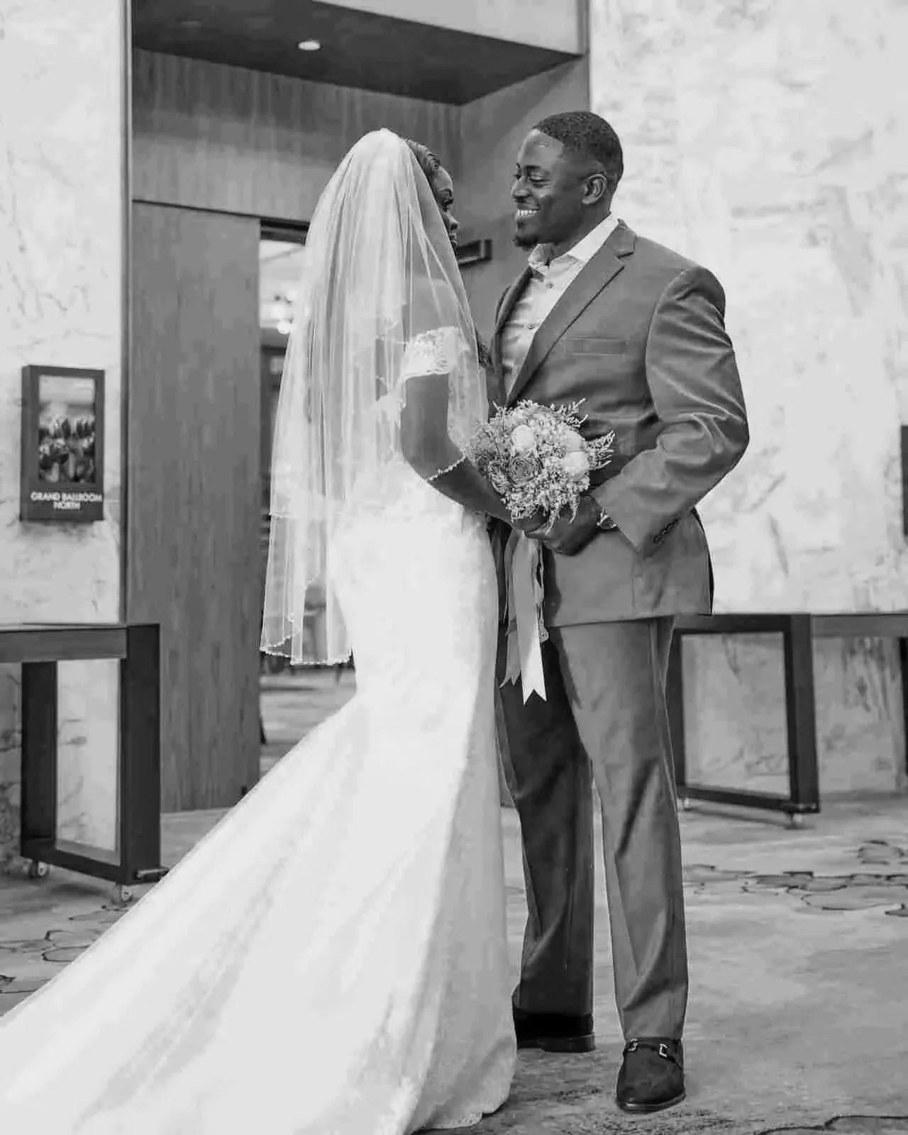 A man with dark curly hair, glasses, and a beard wearing a black knit sweater, sitting indoors near a large green plant.