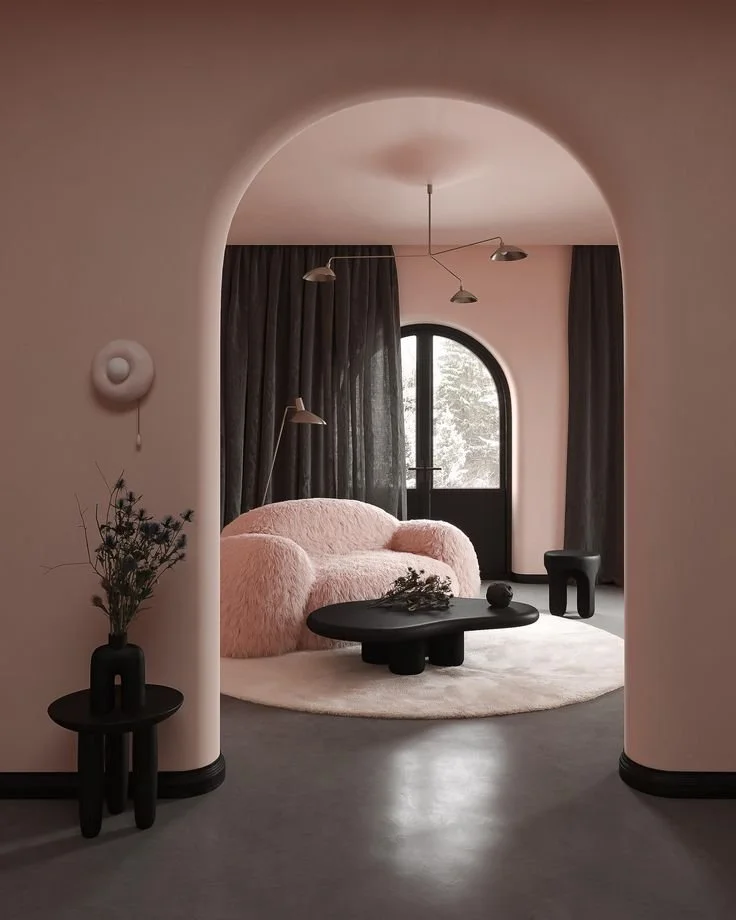 A modern living room with a pink plush sofa, black coffee table, dark curtains, and a large arched window showing a snowy outdoor scene. There are decorative plants and minimalistic lighting fixtures.