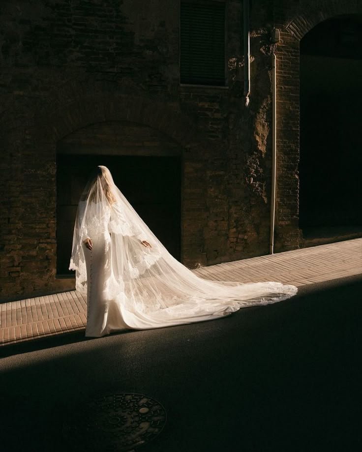 A woman in a wedding dress with a long train, standing on a sidewalk in front of a brick building, with veil covering her face, illuminated by sunlight.