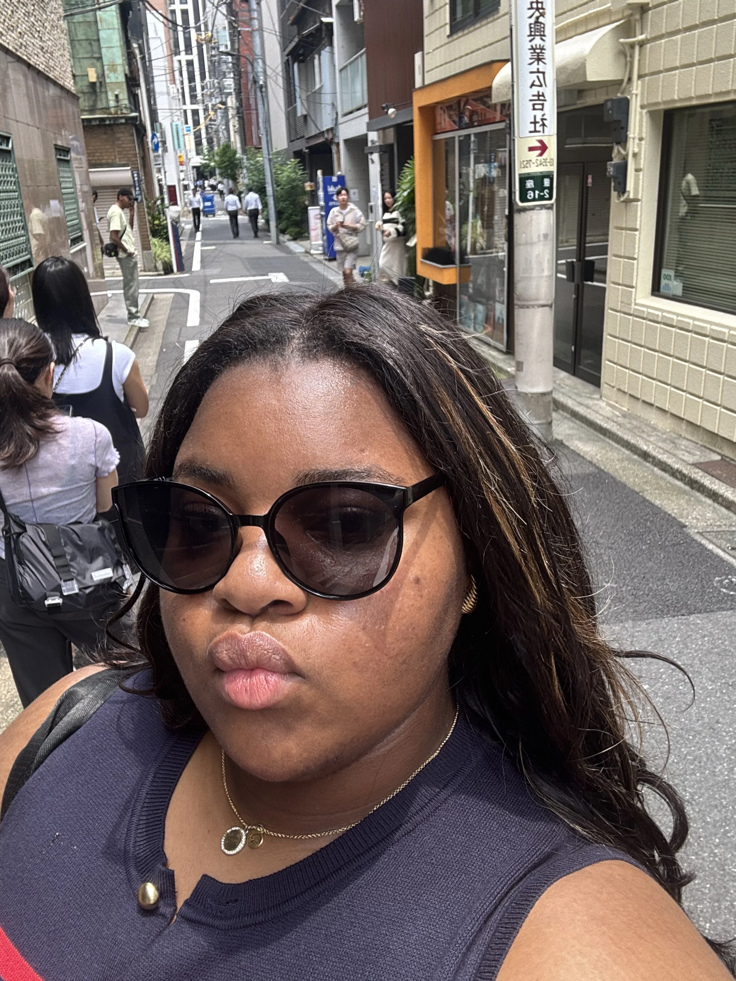 Close-up of a smiling young woman with short, curly hair, glasses, earrings, and jewelry, in an indoor setting.