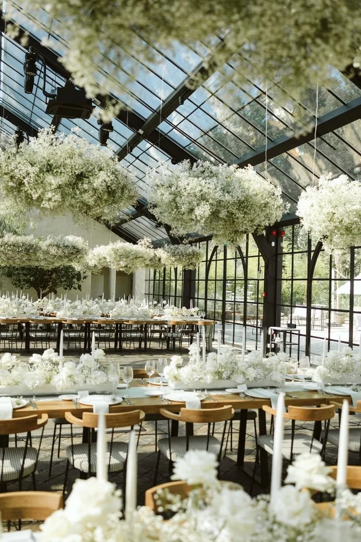 A modern wooden house with a sloped roof sits in a field of tall grass, with a lake and mountains in the background under a cloudy sky.