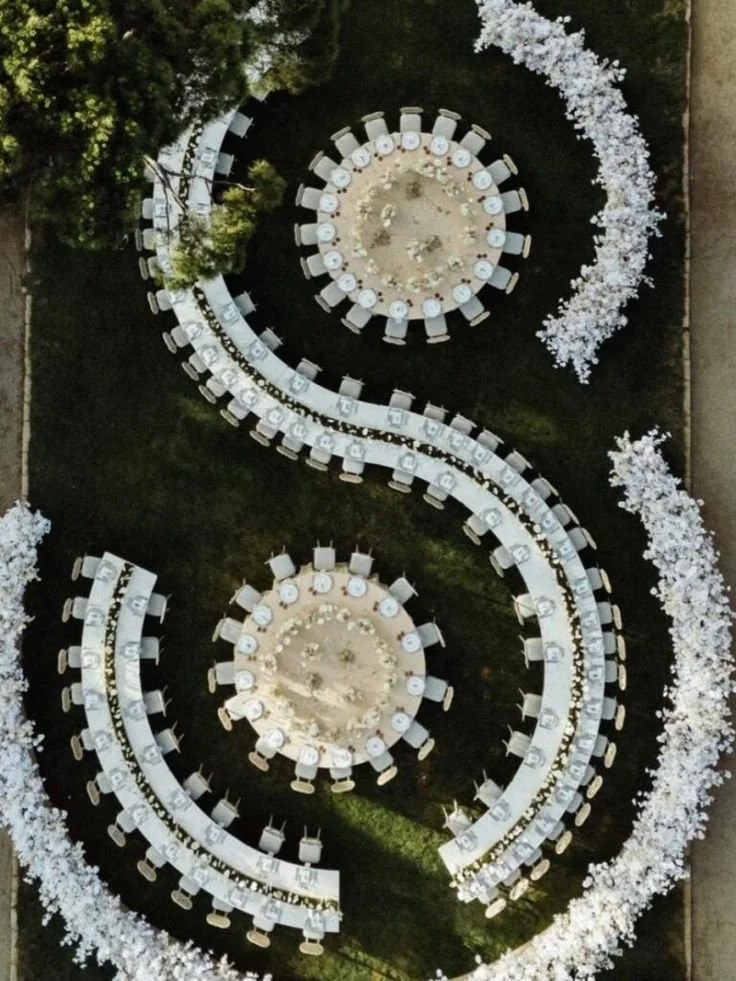 Overhead view of an outdoor event setup with two circular tables, six oval-shaped tables, and a winding table, all decorated with white tableware, surrounded by white floral arrangements.