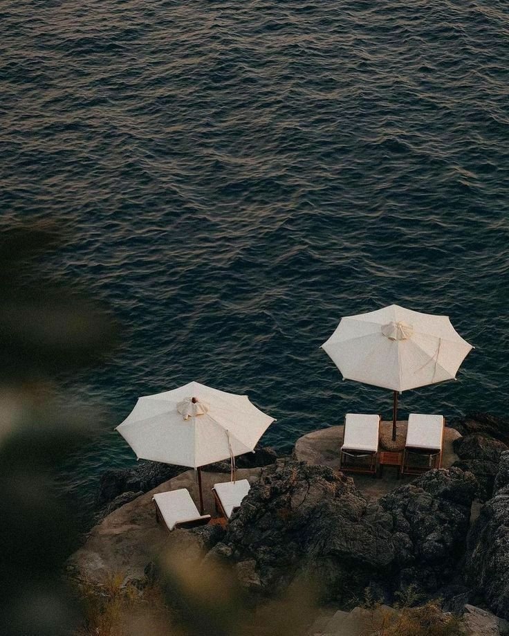 Two white beach umbrellas with lounge chairs on a rocky coastline by the ocean.