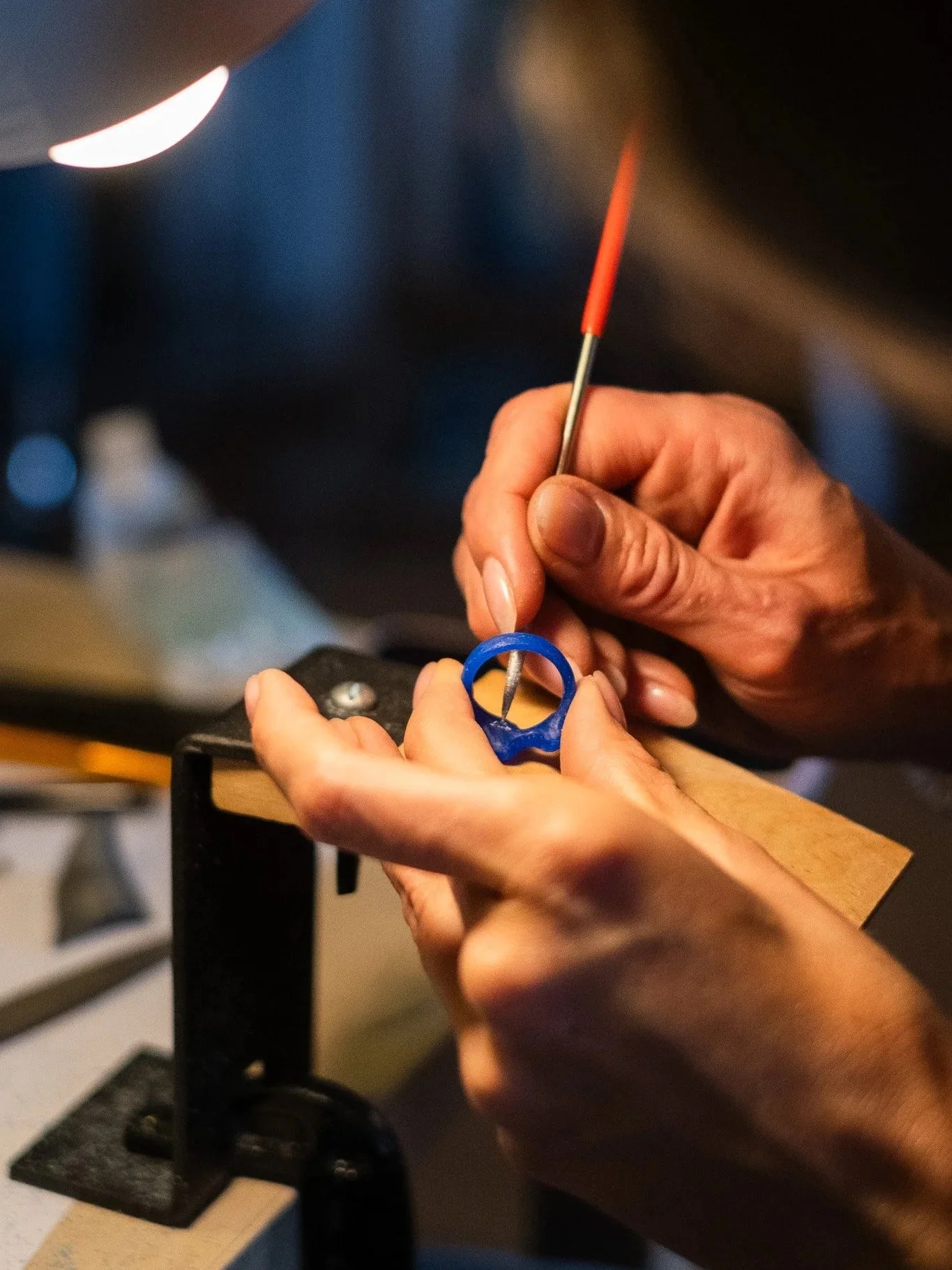 A person using a small file to engrave a blue wax ring during a wax jewelry design workshop. The ring is held in their hand over a black and wooden workbench, with a mount for holding objects in place.