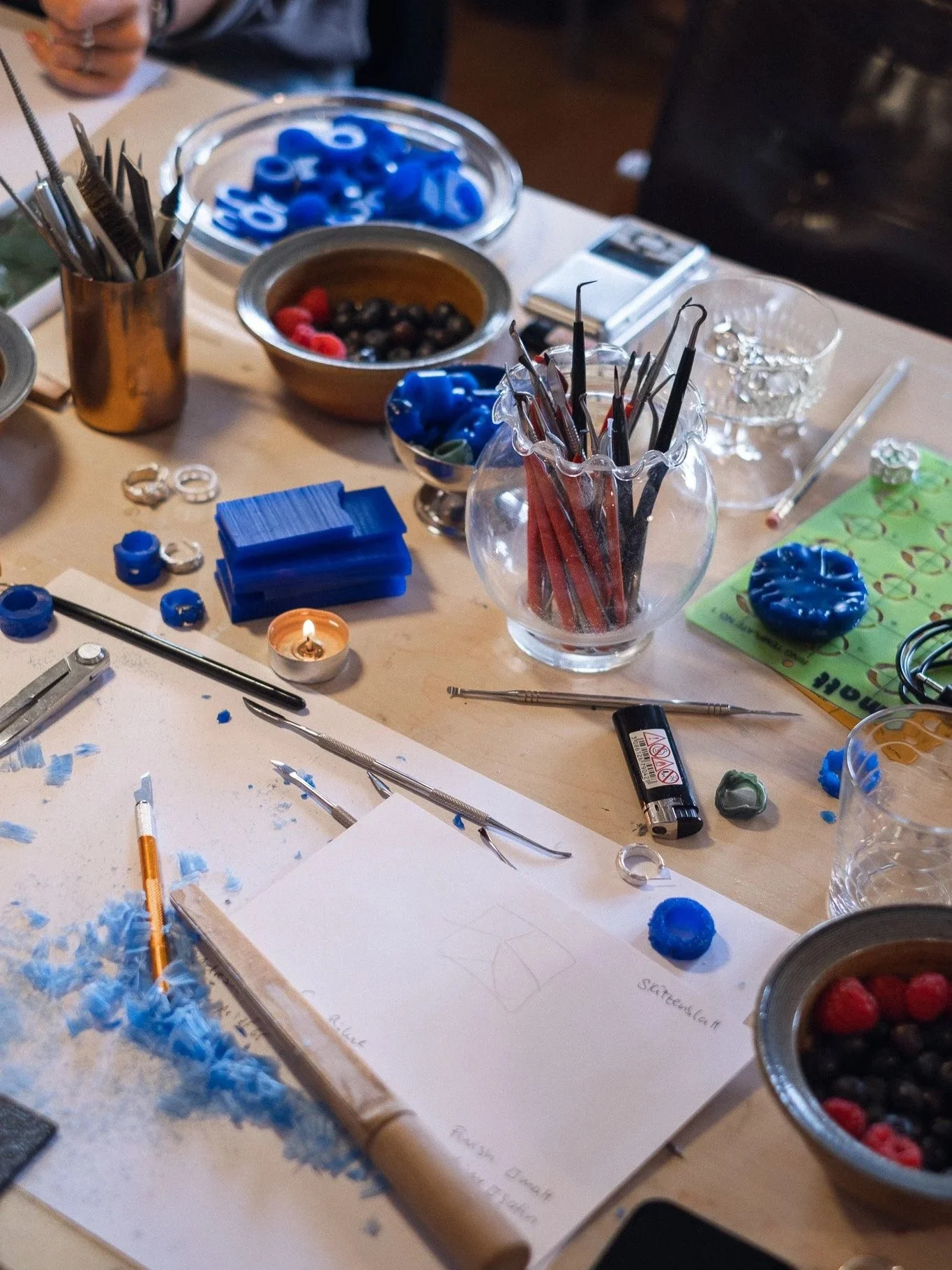 Table with various tools, papers, and materials including blue modelling wax, a candle, a bowl of mixed berries, wax carving tools, drawing material and different types of jewellers wax. Set up for a wax and silver DIY jewelry workshop.
