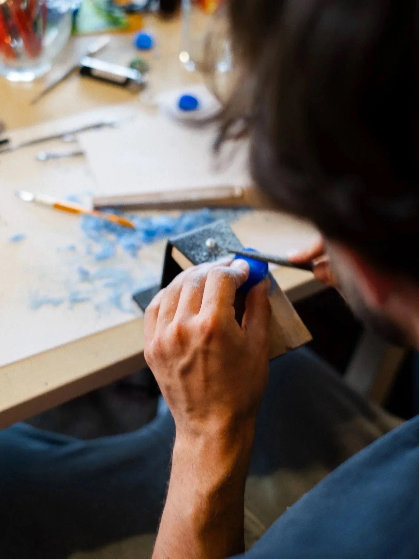 A person using a file to shape a blue wax model ring during a wax jewelry design workshop, sitting at a cluttered workbench with tools and materials.