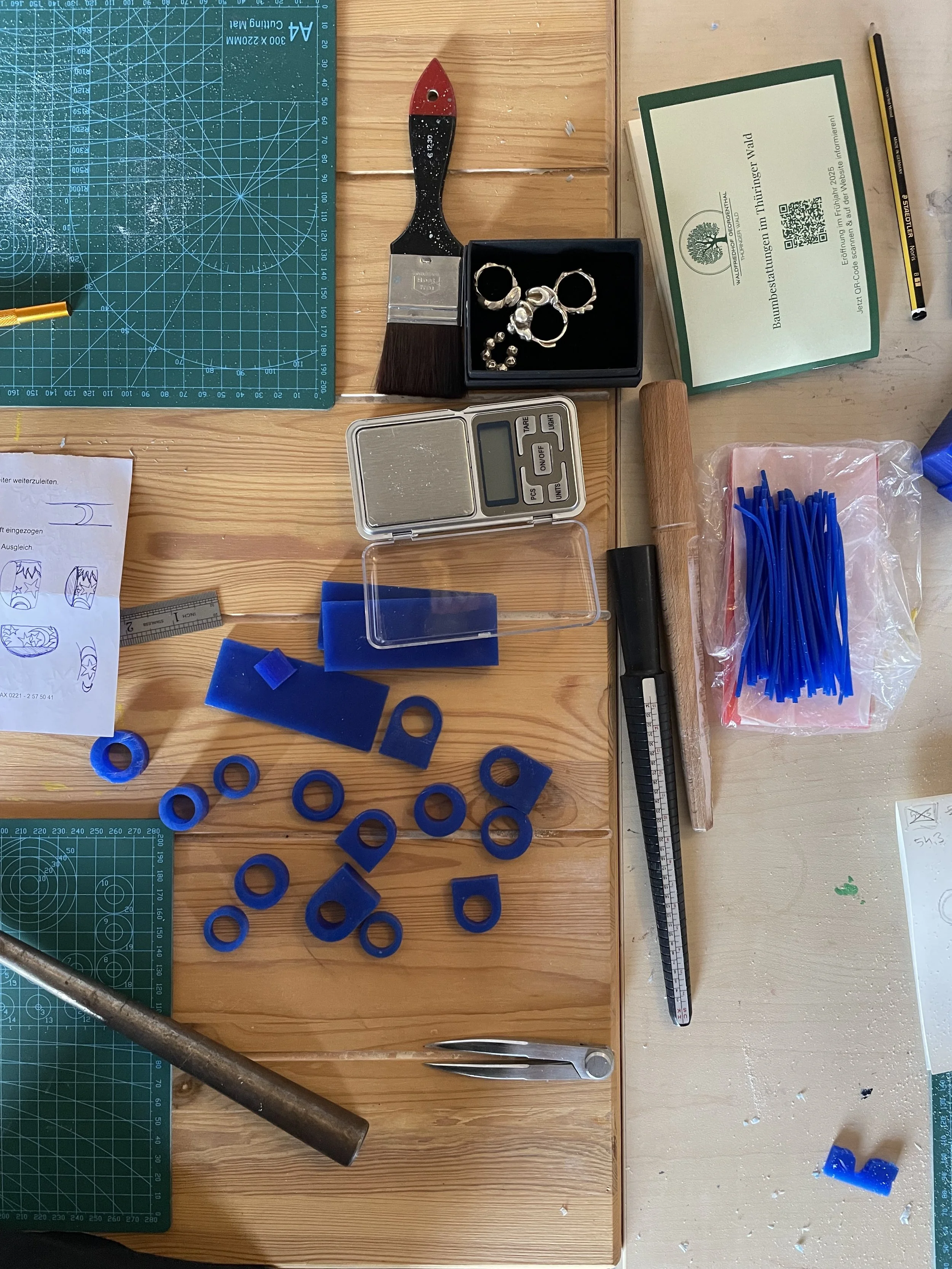 DIY lost wax casting jewelry workshop in Cologne with jewelry-making tools and supplies on a wooden workspace, including a digital scale, blue wax rings, pliers, a hammer, a cutting mat, a ruler, jewelry findings, and a box with silver rings.
