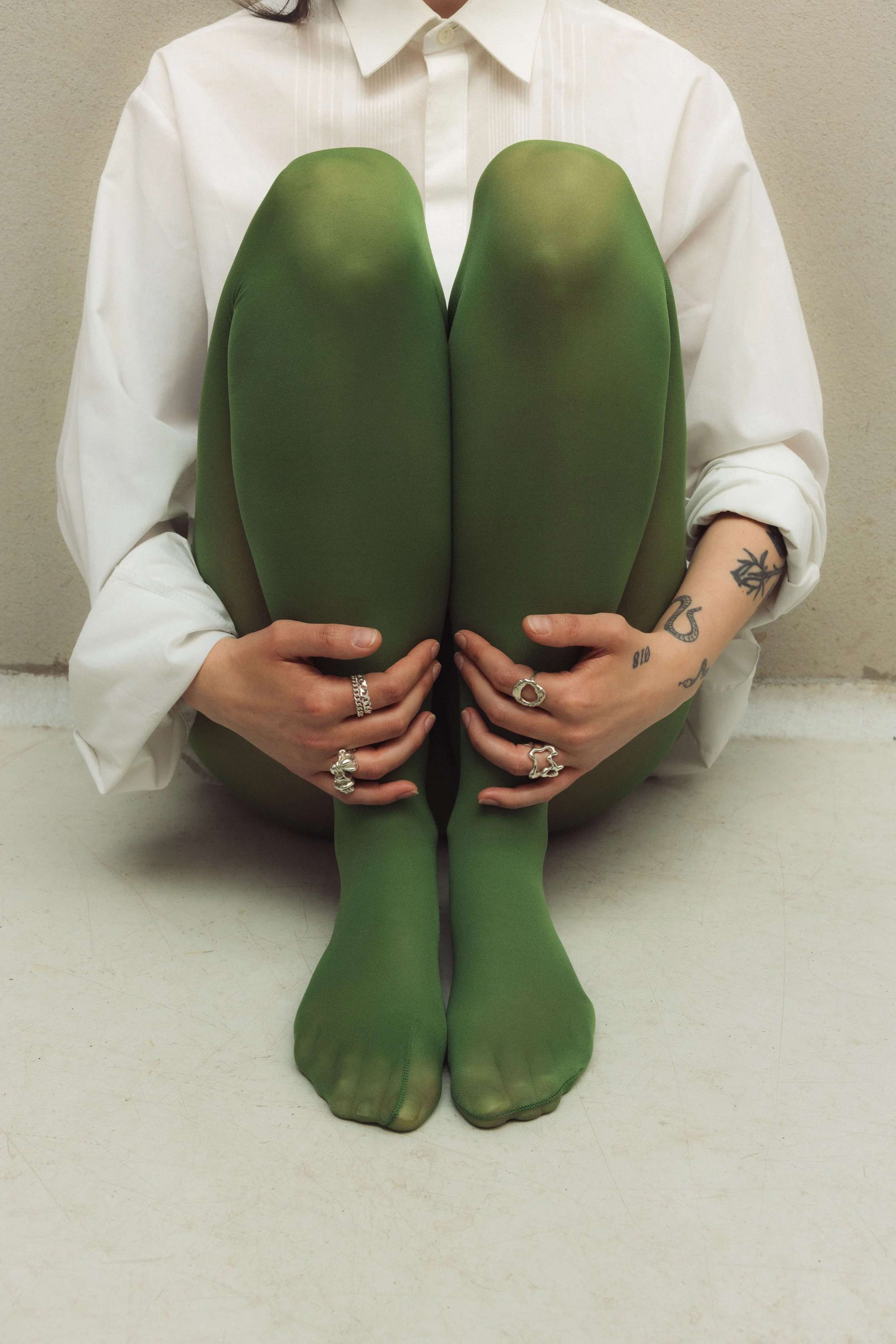 Person sitting on the floor wearing a white shirt, green tights, and multiple unique 925 silver rings, holding their legs in front of their chest.