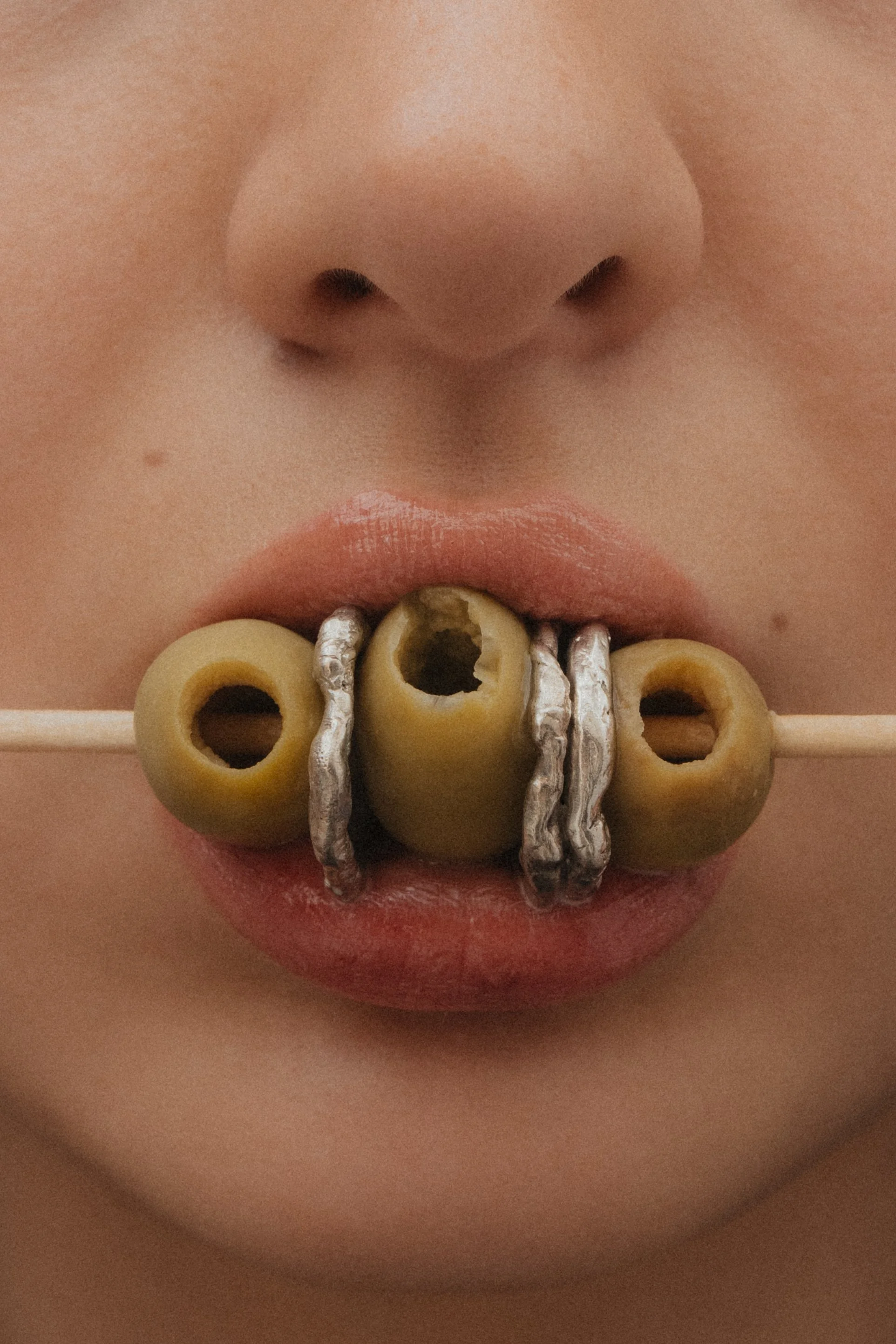 Close-up of a person's lips with olives on a stick, textured silver rings stacked between them, held in their mouth.