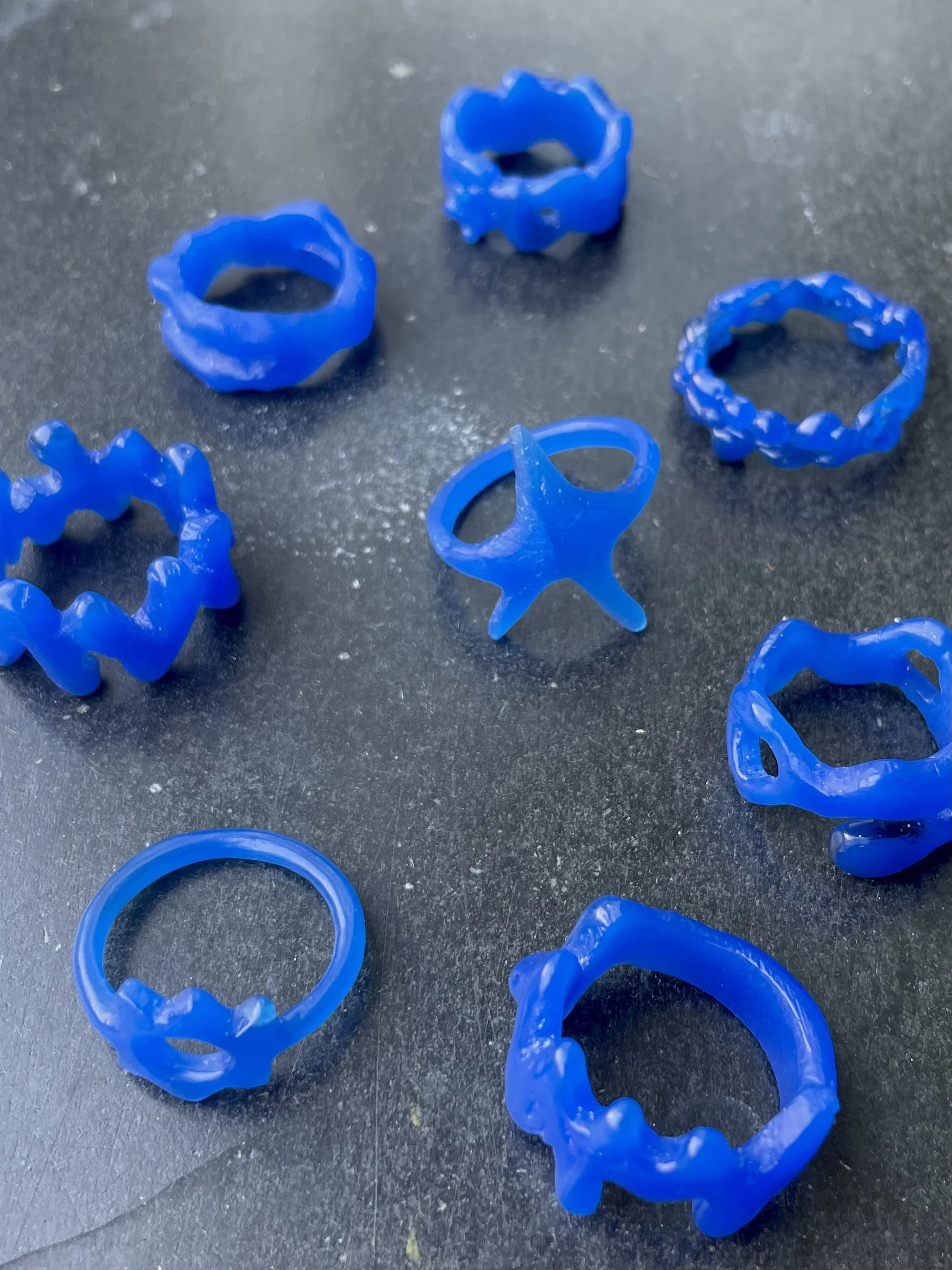 Several blue wax rings with various textured and shaped designs scattered on a black surface that are the result of a lost wax casting jewelry workshop in Köln.