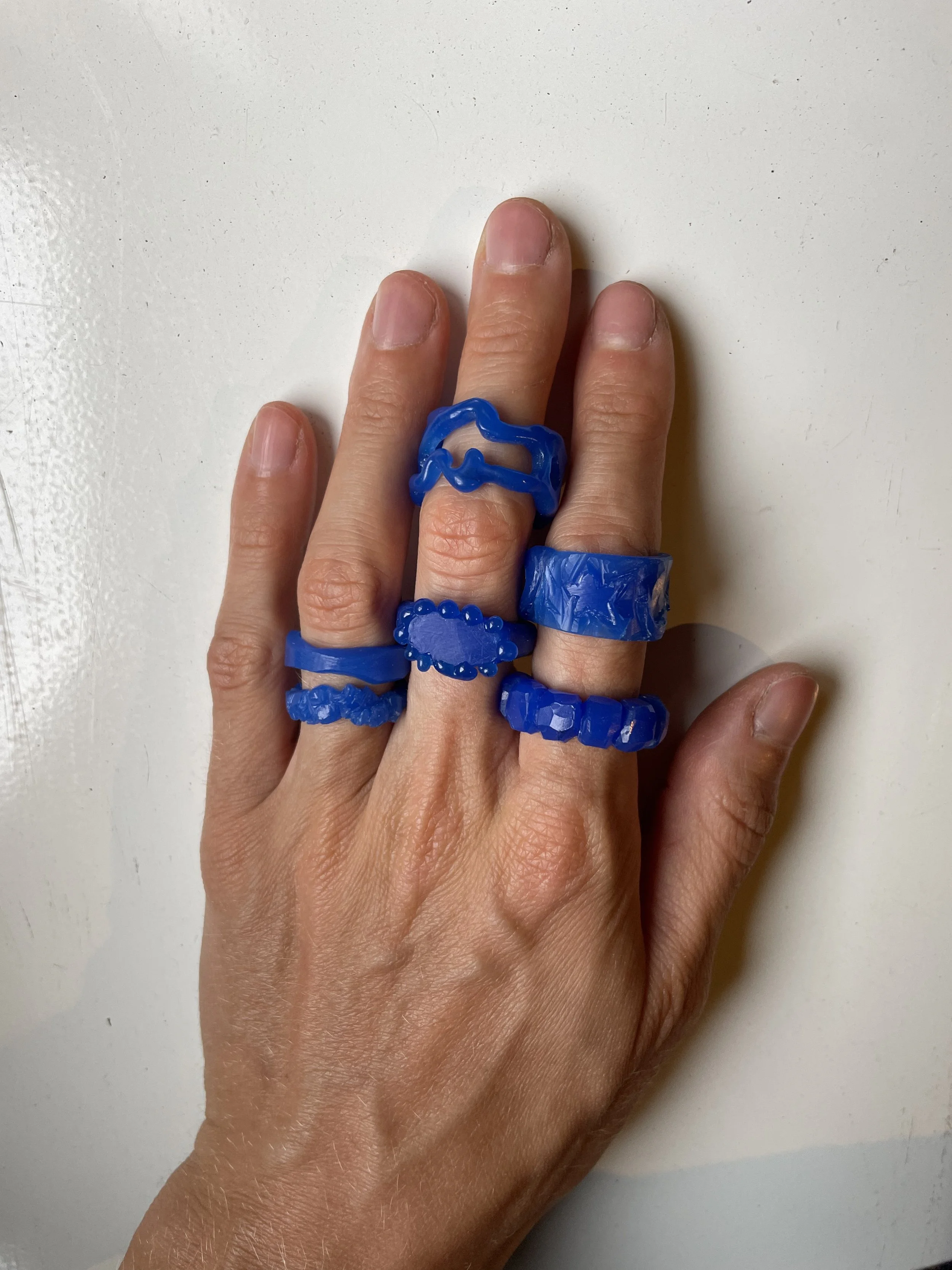 Hand wearing multiple blue wax rings with various textured and shaped designs that are the result of a lost wax casting jewelry workshop in Köln, resting on a white surface.