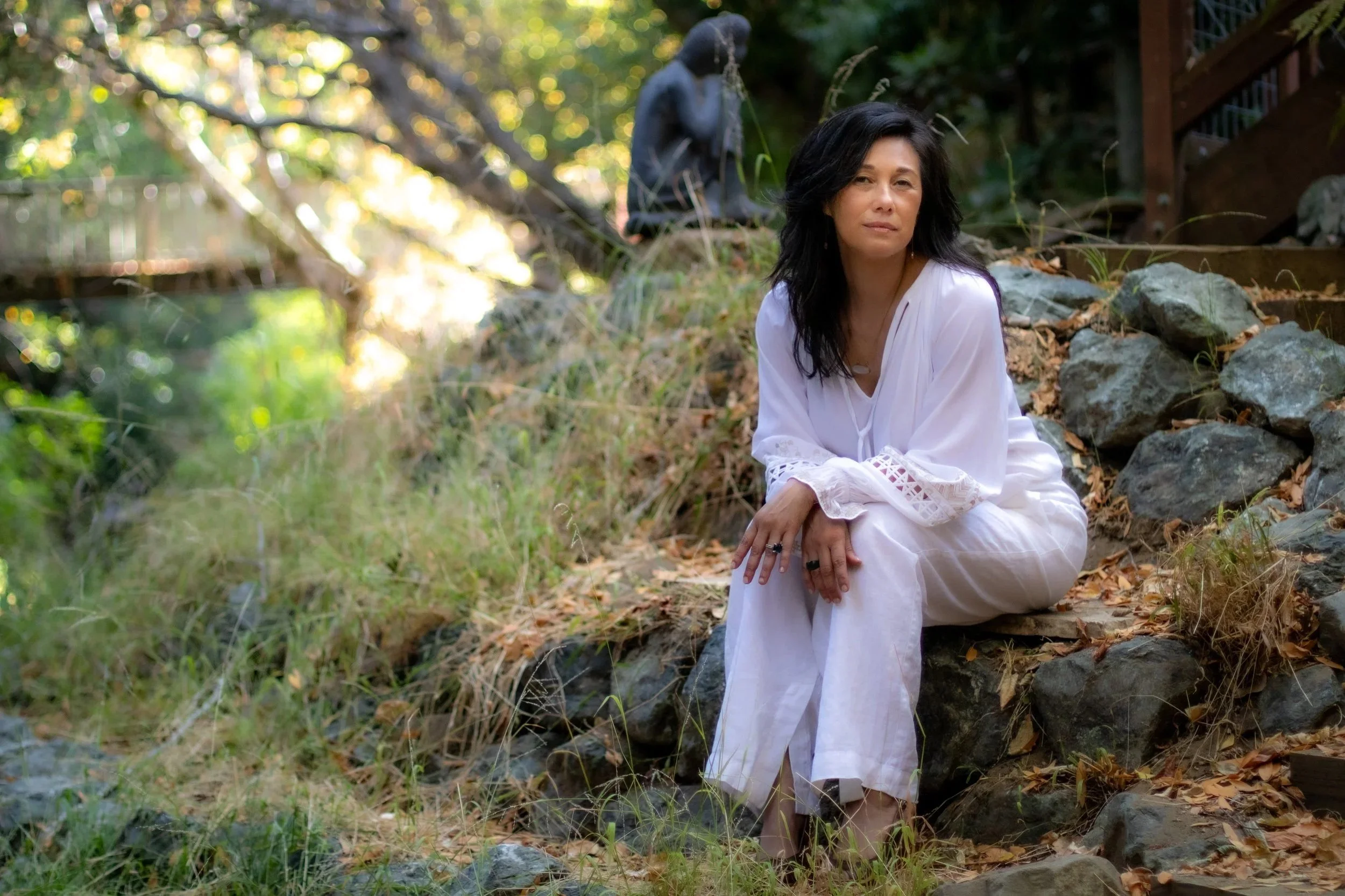A woman in a white dress sitting on rocks outdoors with trees and a statue in the background.