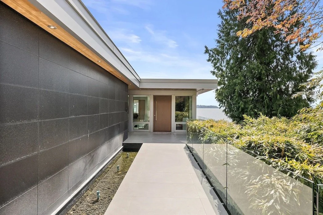 Modern building entrance with a walkway, glass railing, trees, and a body of water in the background.