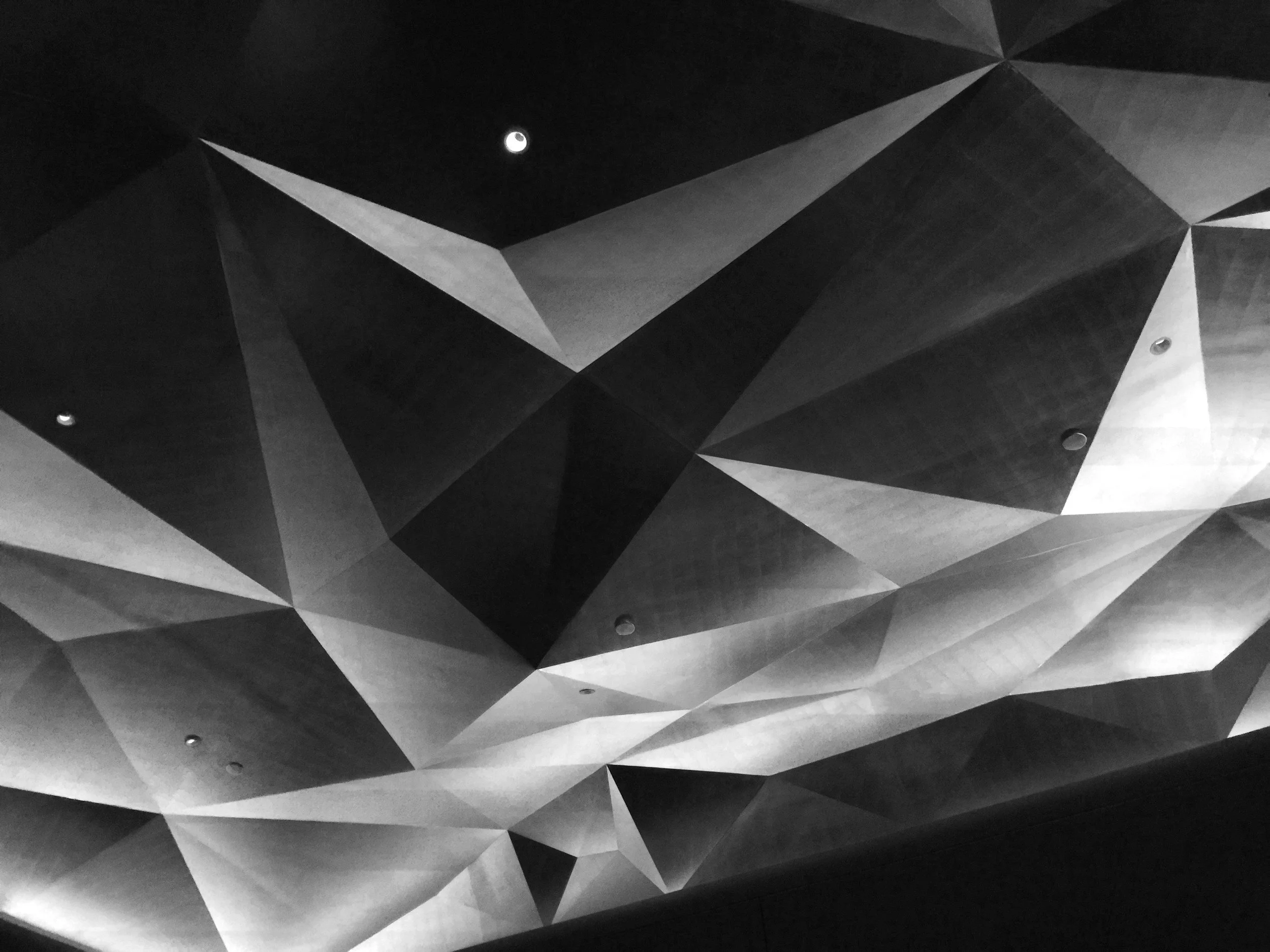 Abstract geometric ceiling with various triangular shapes, lighting fixtures, and a visible moon through a small opening.