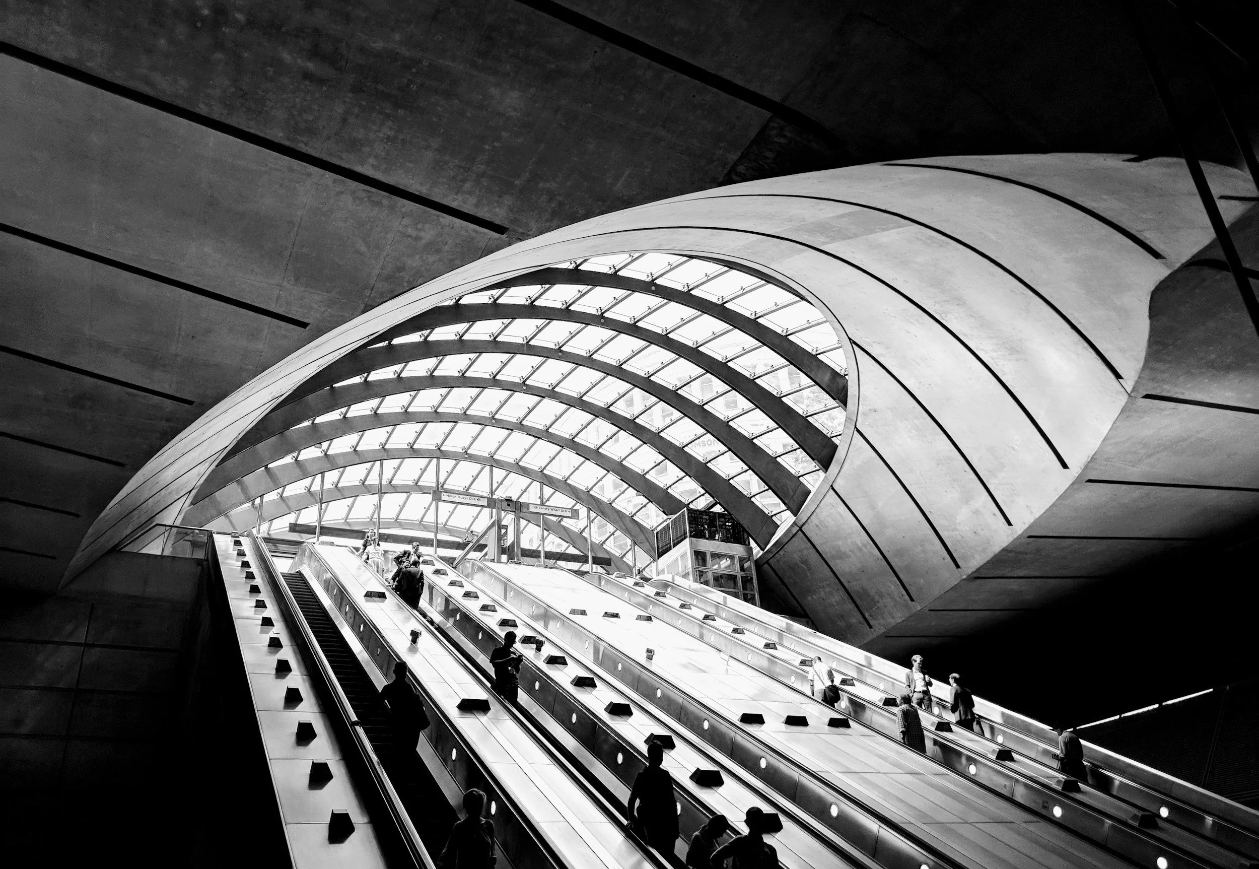 Black and white photo of an underground train station with a modern, curved glass ceiling and escalators with people ascending and descending.