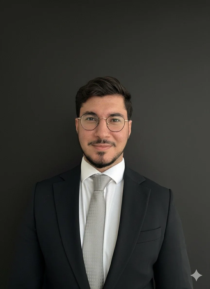 A young man with dark hair and glasses is wearing a black suit, white shirt, and silver tie, standing in front of a plain dark background.
