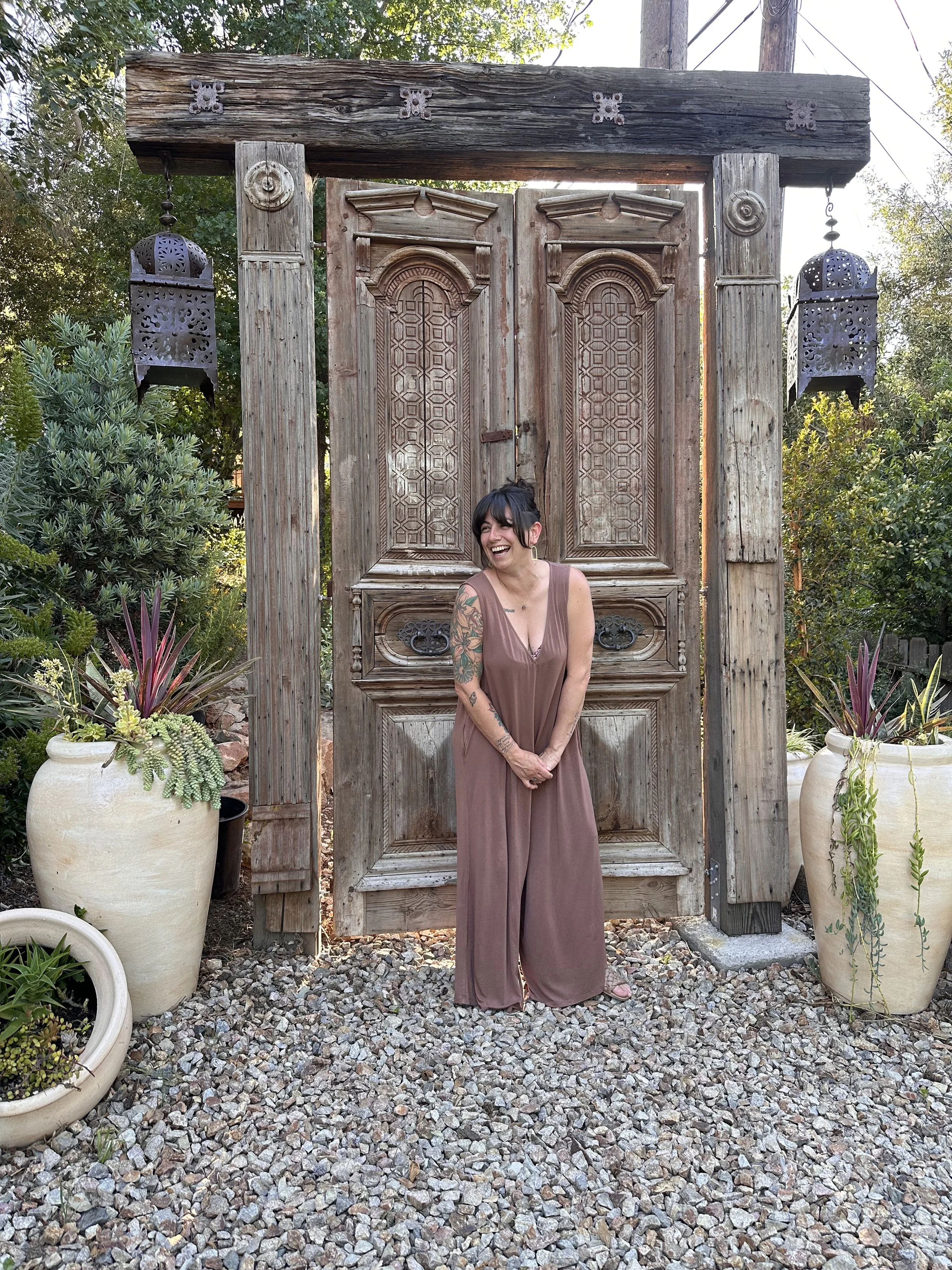 A woman with black hair and tattoos on her arms, smiling and wearing a brown jumpsuit, standing in front of an ornate, rustic wooden door surrounded by potted plants.