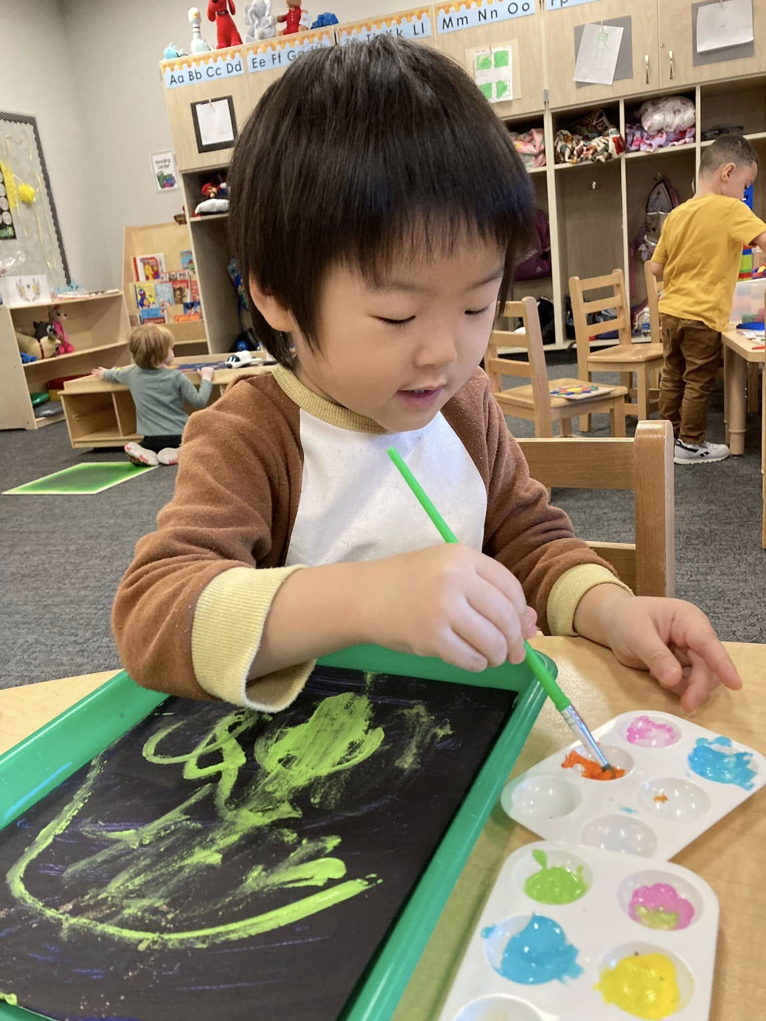 Young boy with dark hair drawing on a black chalkboard with green paint, sitting at a small table in a classroom with other children and shelves of educational materials in the background.