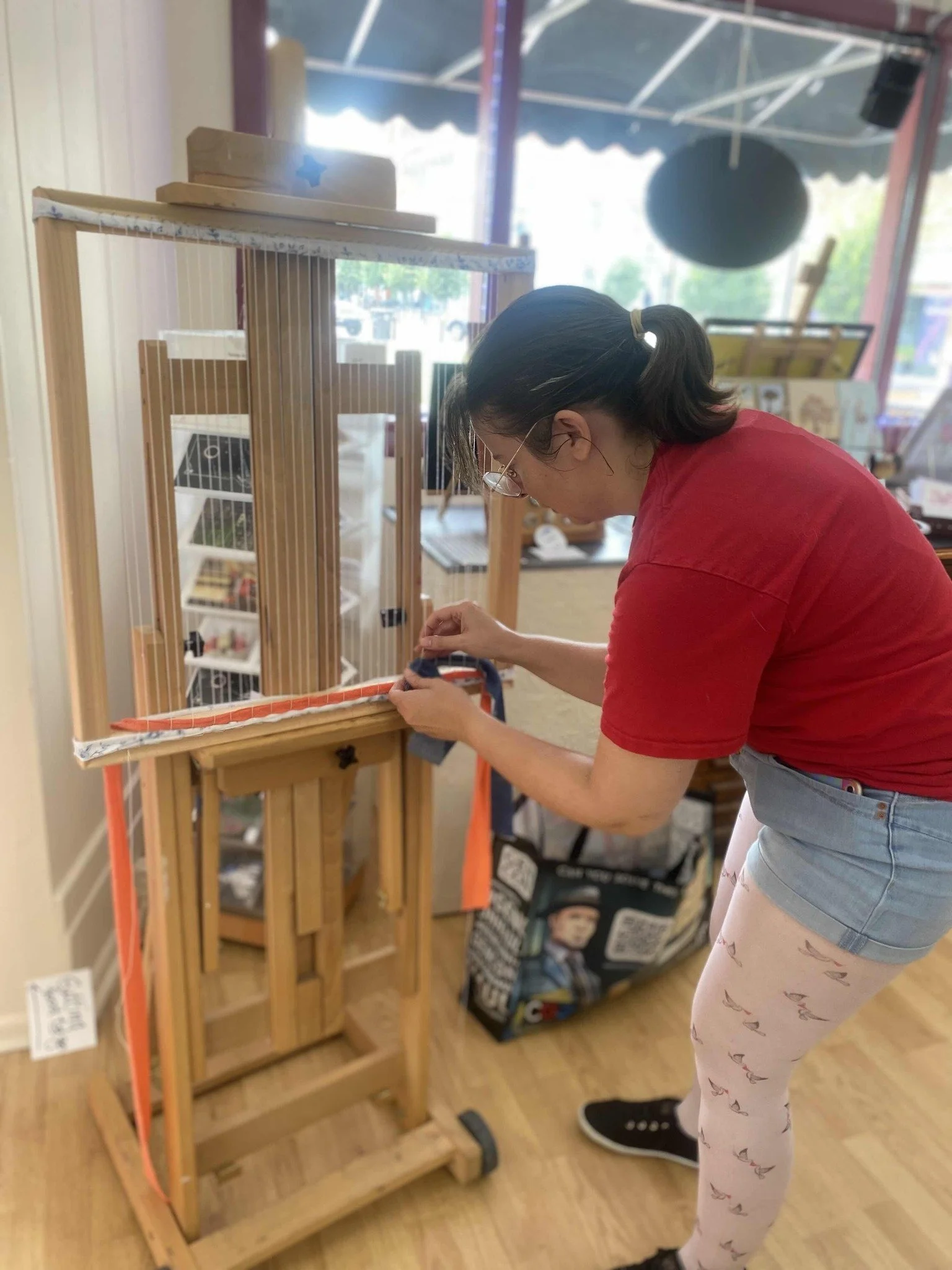 A woman with glasses, wearing a red t-shirt, denim shorts, and pink leggings with flying birds, is tying a string on a small wooden puppet theater display inside a store.