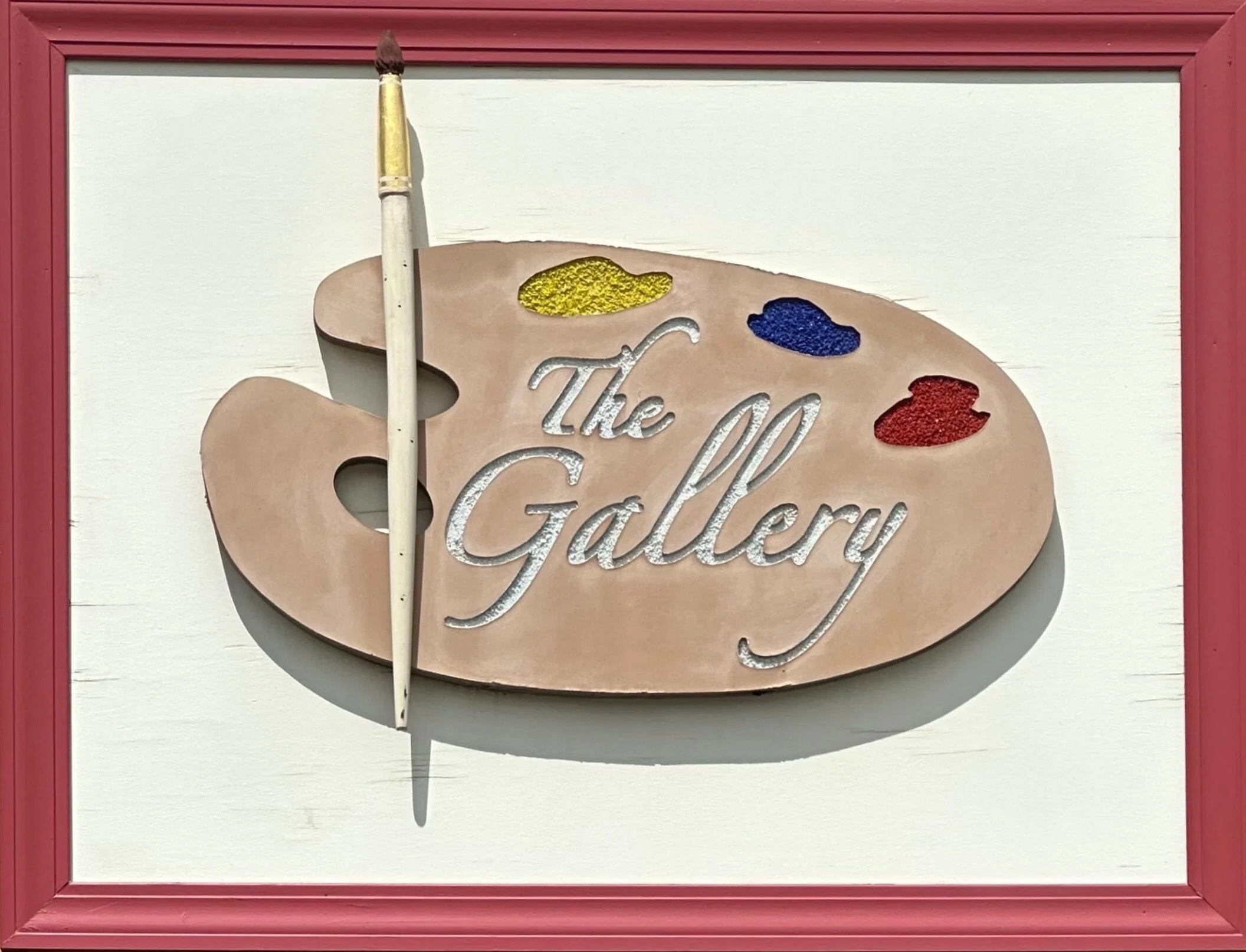 Sign for an art gallery featuring a painter's palette with splashes of yellow, blue, and red paint, and the words 'The Gallery' engraved on the palette. A paintbrush is attached to the palette.