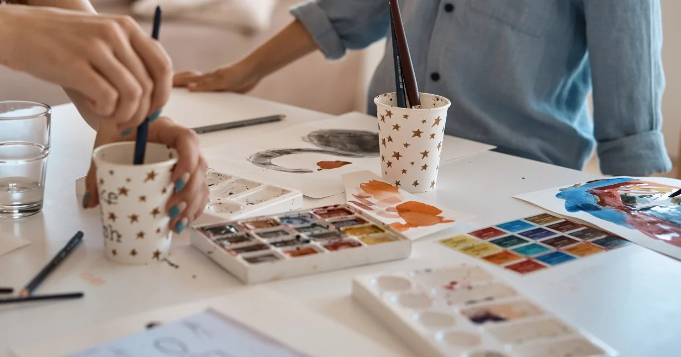 People painting and drawing at a table with watercolor paints and brushes.