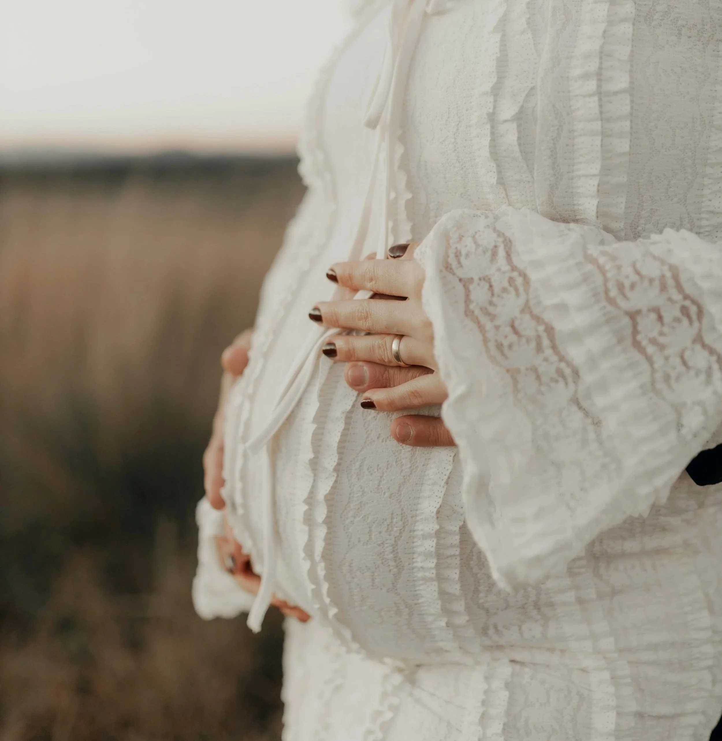 A close-up of a pregnant woman wearing a white lace dress, gently cradling her baby bump with one hand.