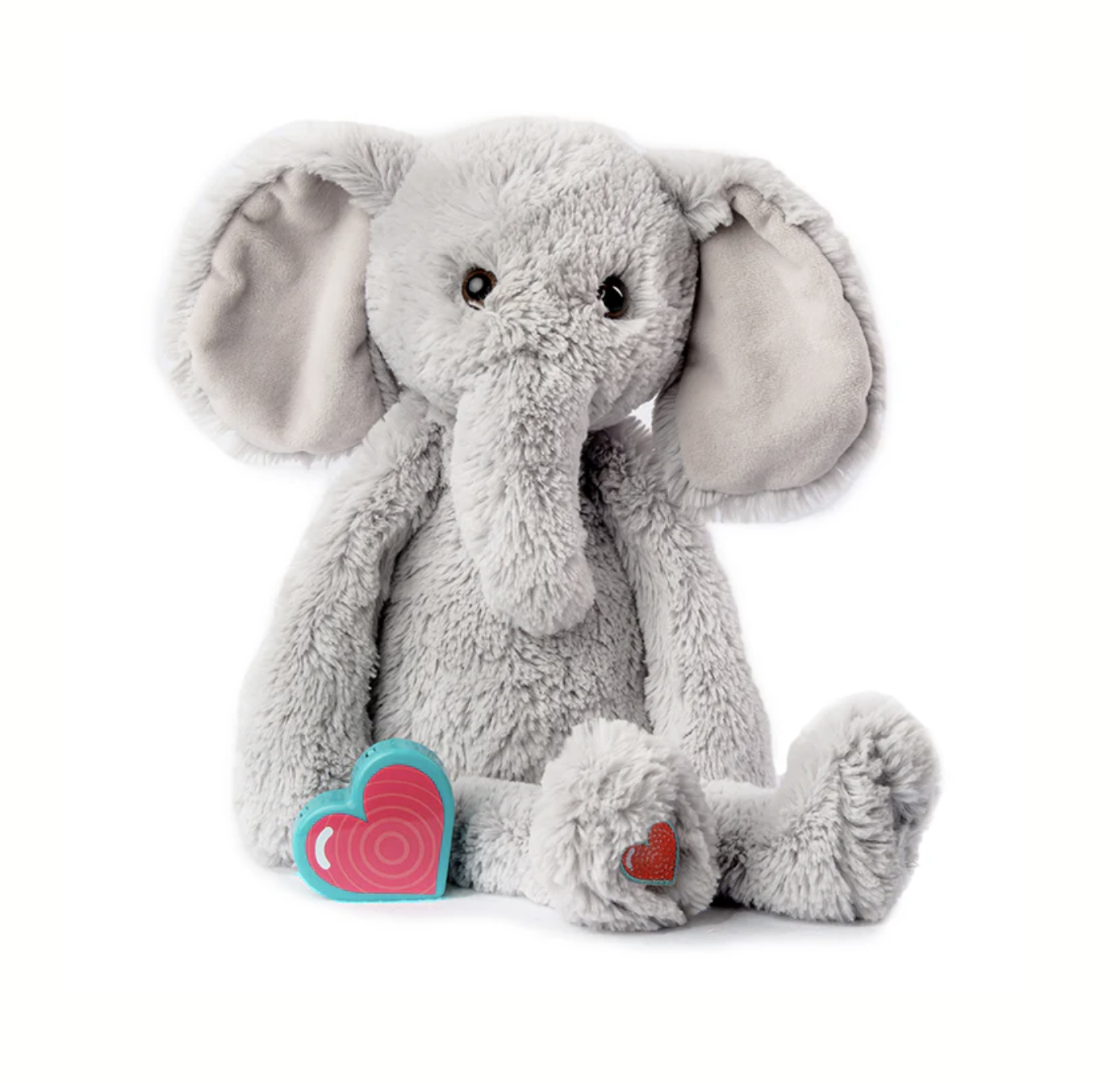 A plush gray elephant stuffed toy with large floppy ears, sitting on a white background with a pink and blue heart-shaped object next to it and a small red heart on its foot.