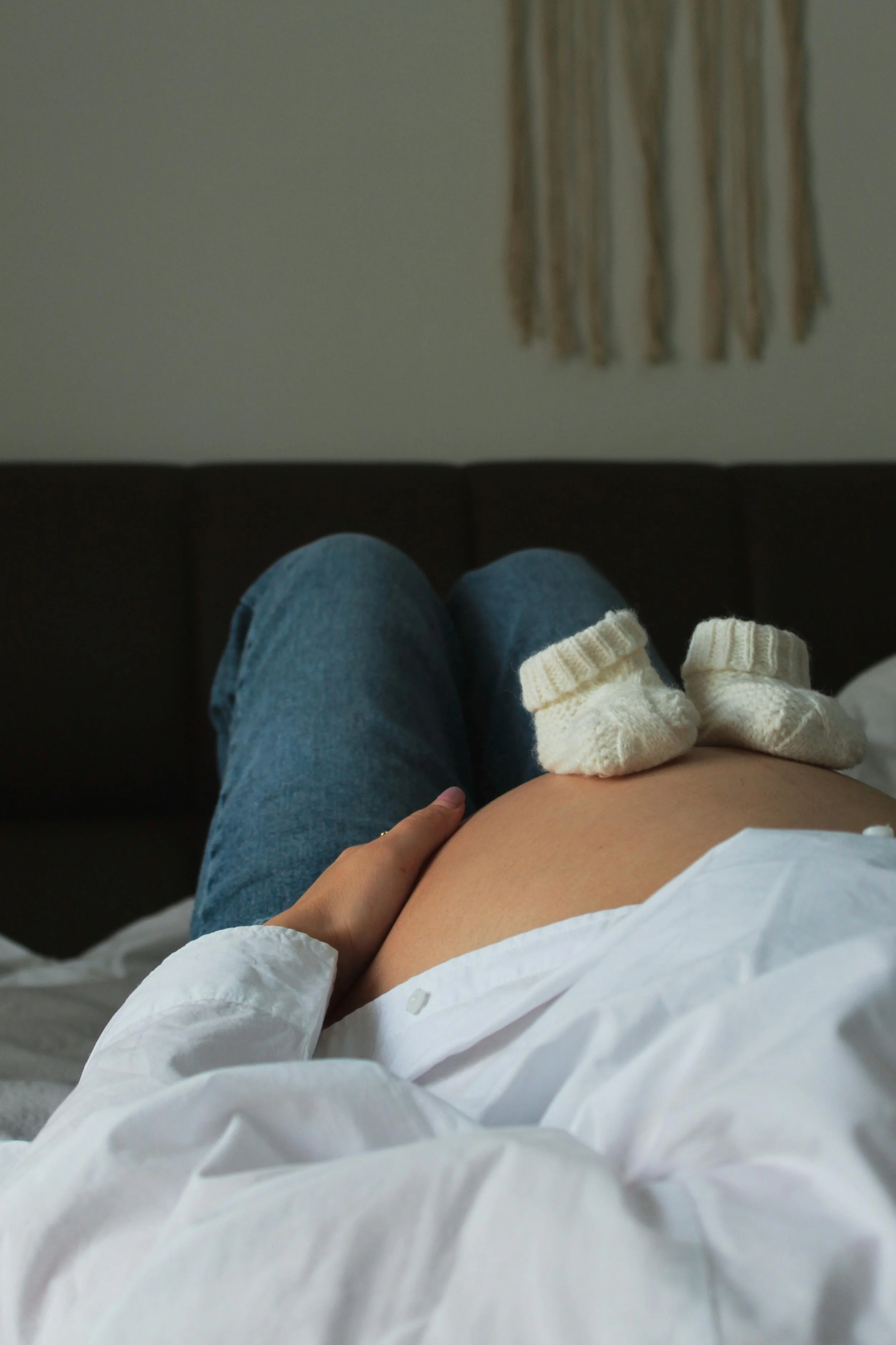 Person lying on bed with legs up, wearing blue jeans and a white shirt, with baby socks on their pregnant belly in a softly lit bedroom.