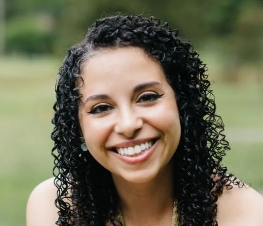 Close-up of a smiling woman with curly black hair outdoors.