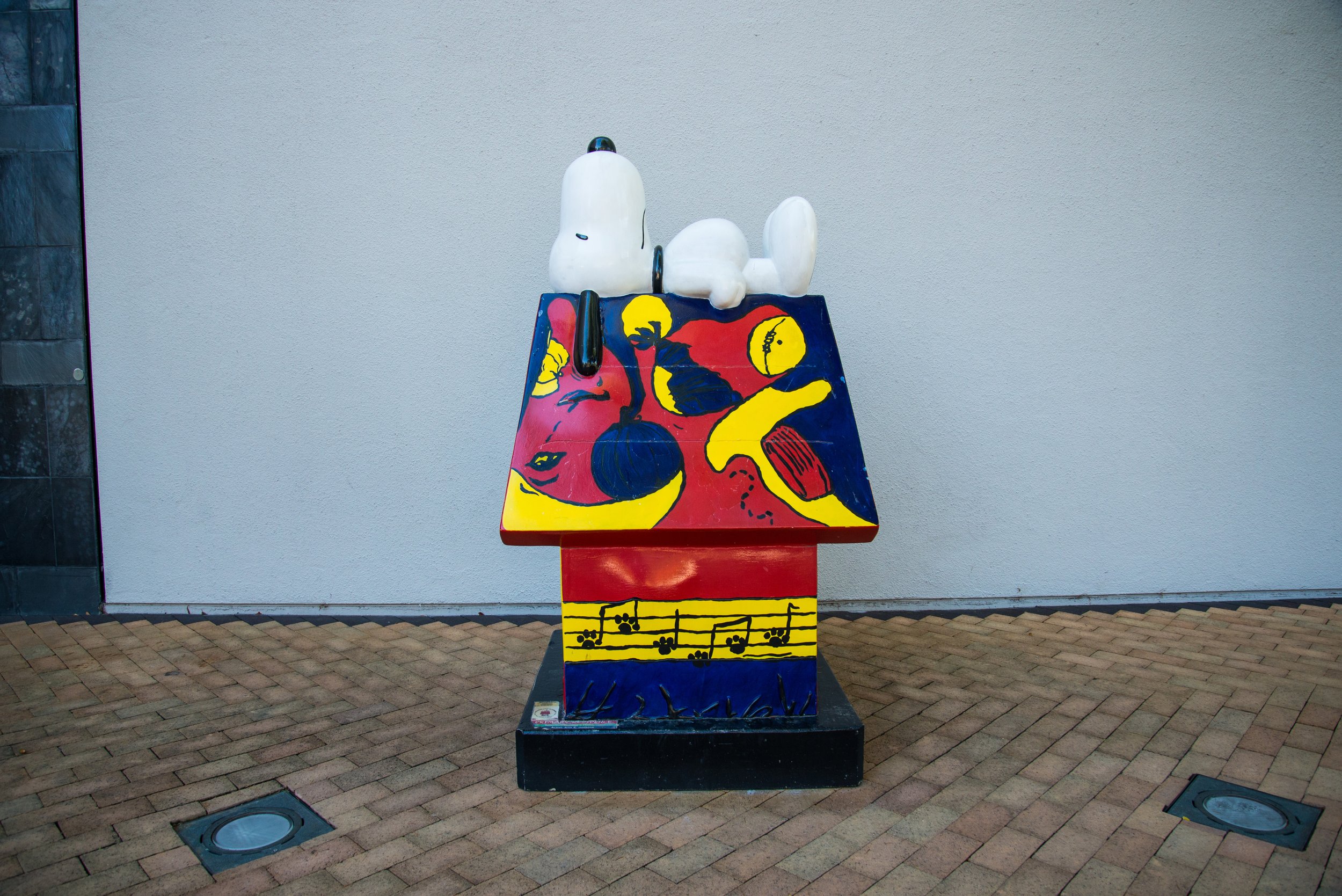 Colorful Snoopy statue lying on painted doghouse with vibrant geometric designs and musical notes, outdoor setting.
