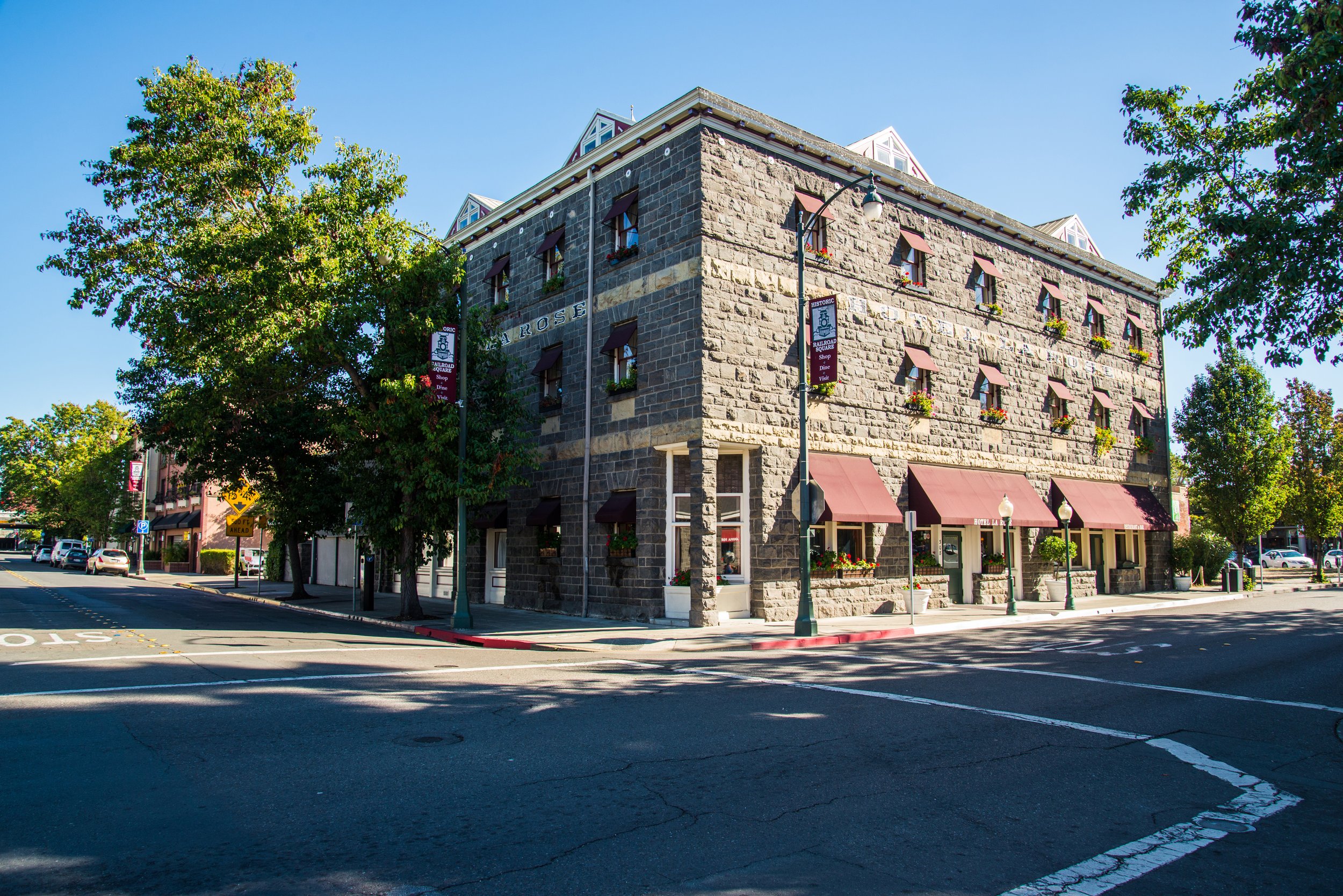 Historic stone hotel building on a sunny street corner with red awnings and potted plants, surrounded by trees and street signage.