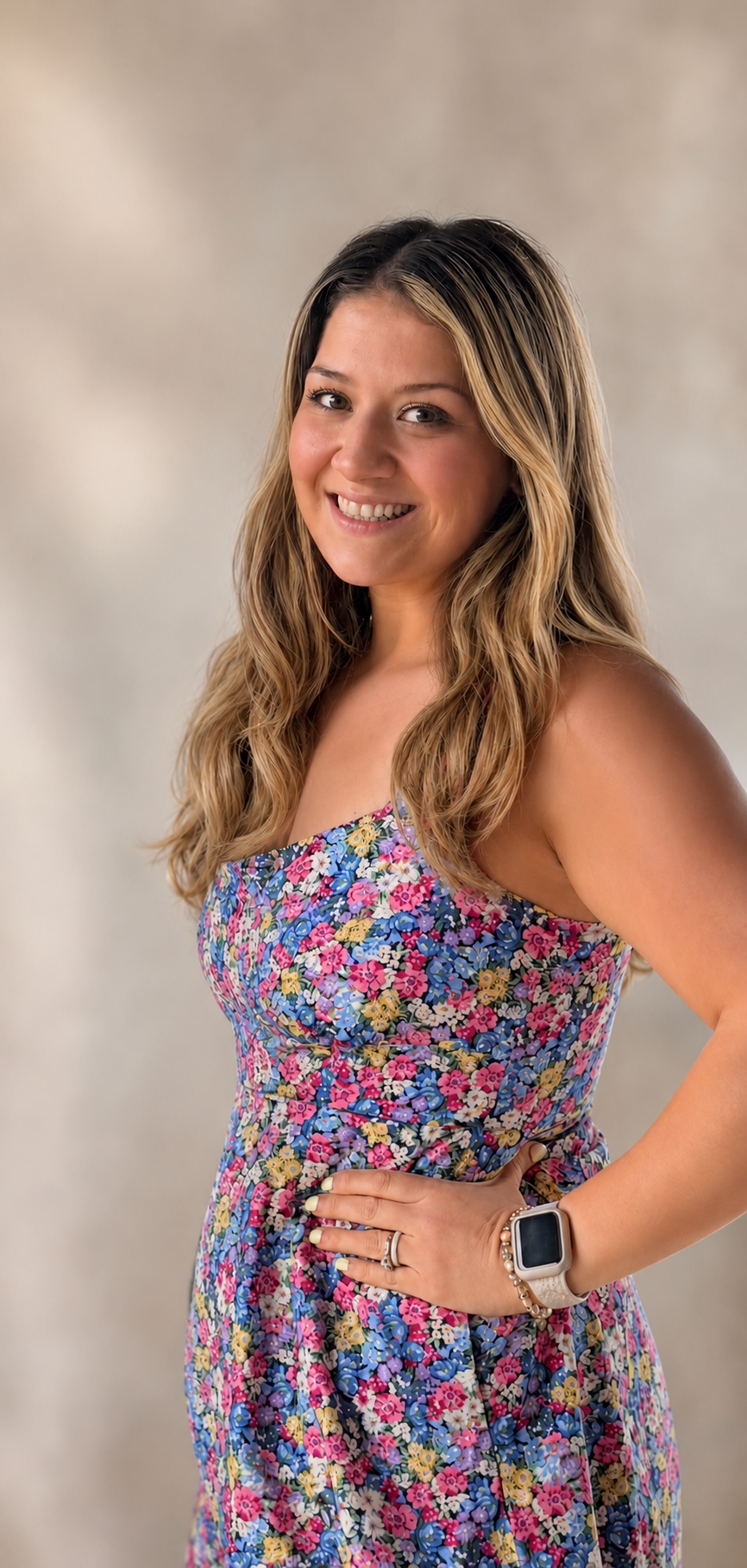 A woman with long, wavy blonde hair wearing a colorful, floral dress and a smartwatch, posing with her hand on her hip and smiling at the camera.