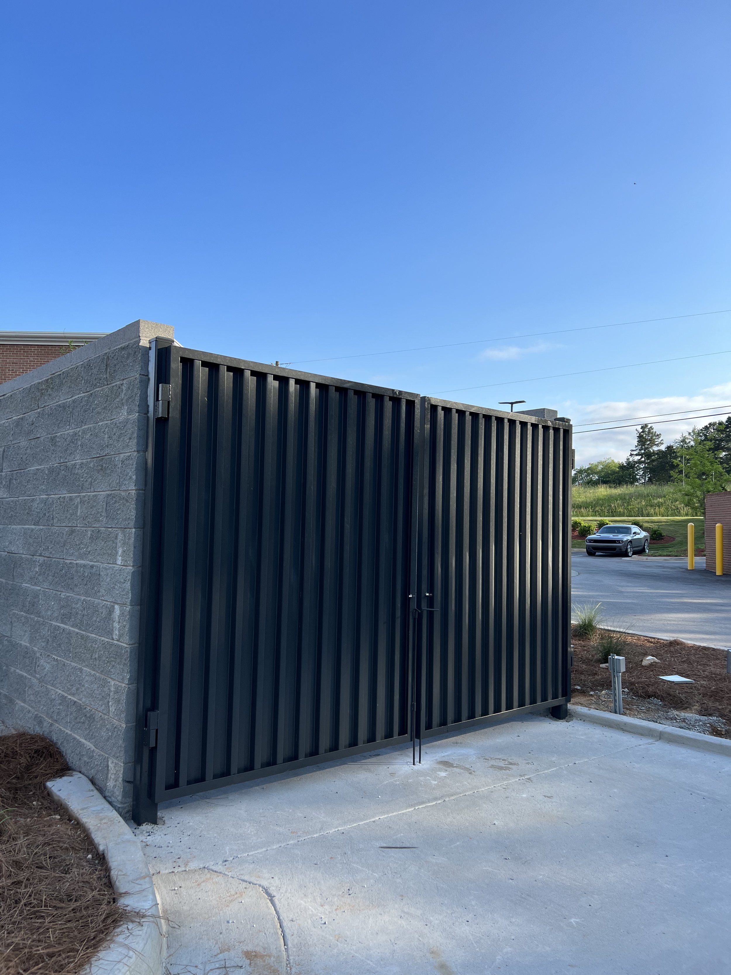 A black metal dumpster enclosure with closed doors next to a brick wall. The area is paved with concrete and there is a clear blue sky in the background.