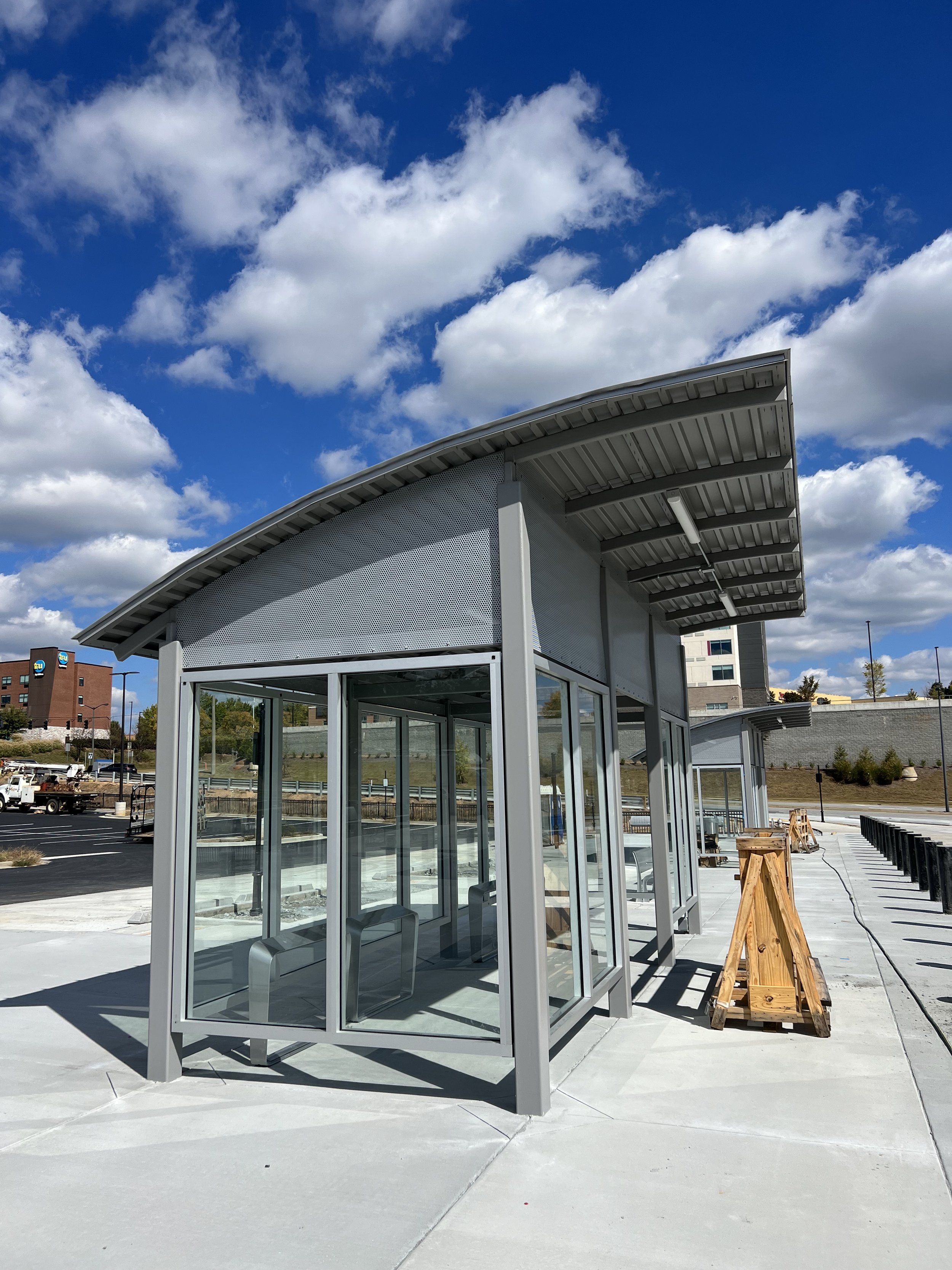 A small glass and metal structure under a curved roof at a construction site, with a blue sky and clouds in the background.