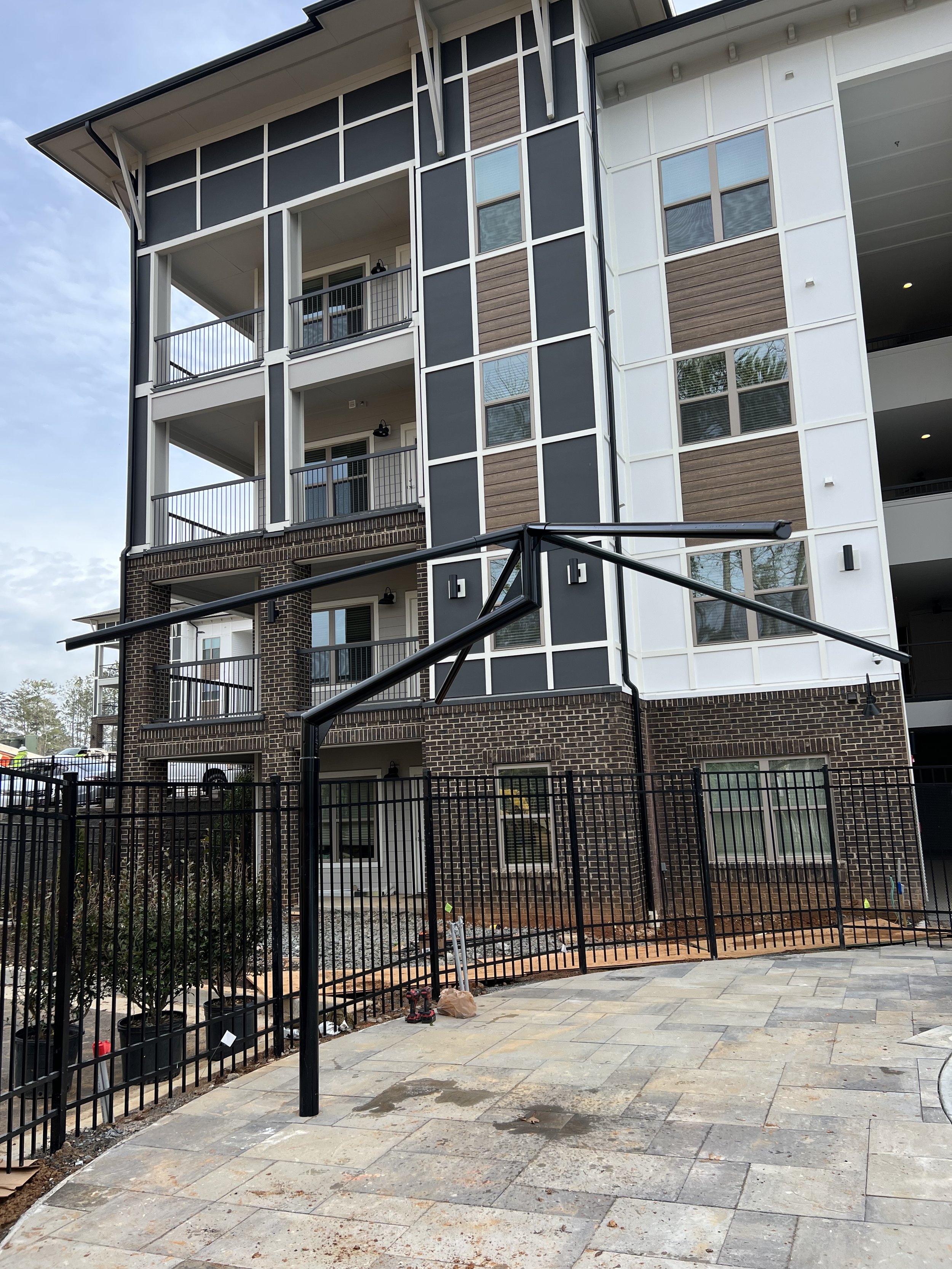 Exterior view of a multi-story residential building with balconies, brick and siding façade, surrounded by a black metal fence and a tiled patio area under construction.