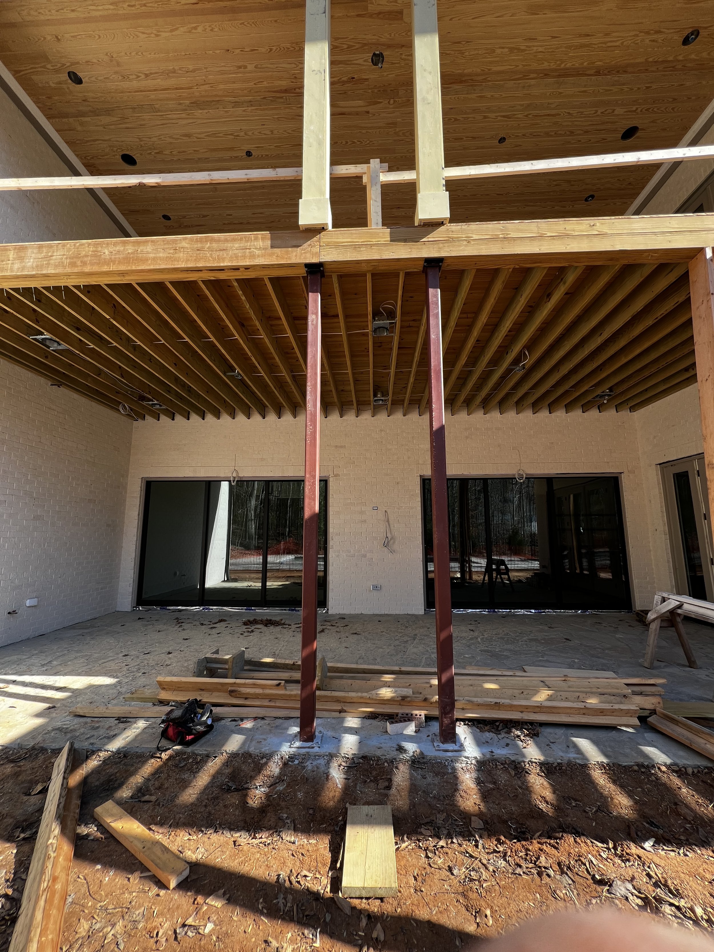 Construction site with unfinished wooden roof structure, metal support beams, and scattered lumber in front of a building with glass doors.