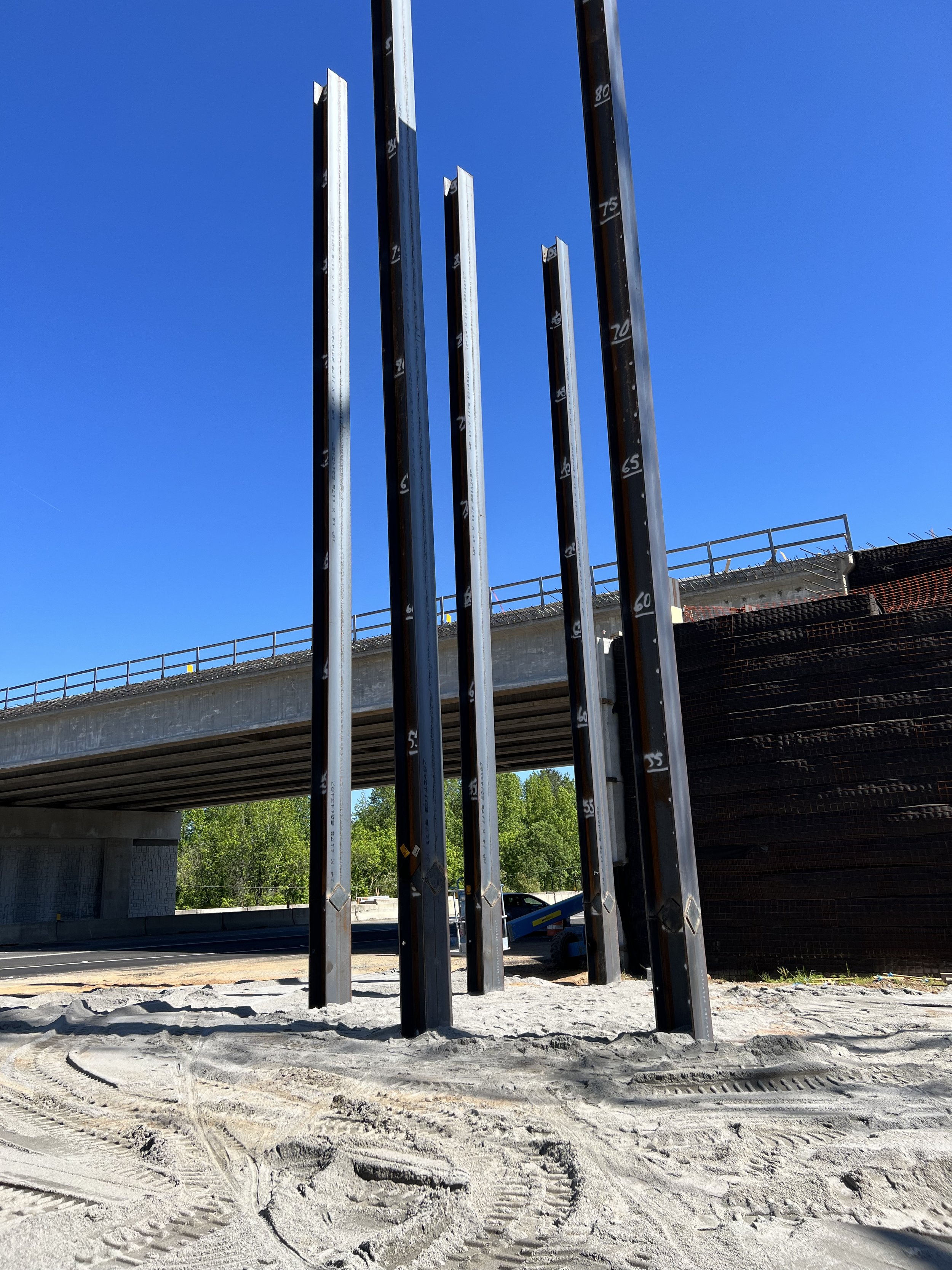 Metal beams installed vertically at a construction site near a road bridge, with a clear blue sky in the background.