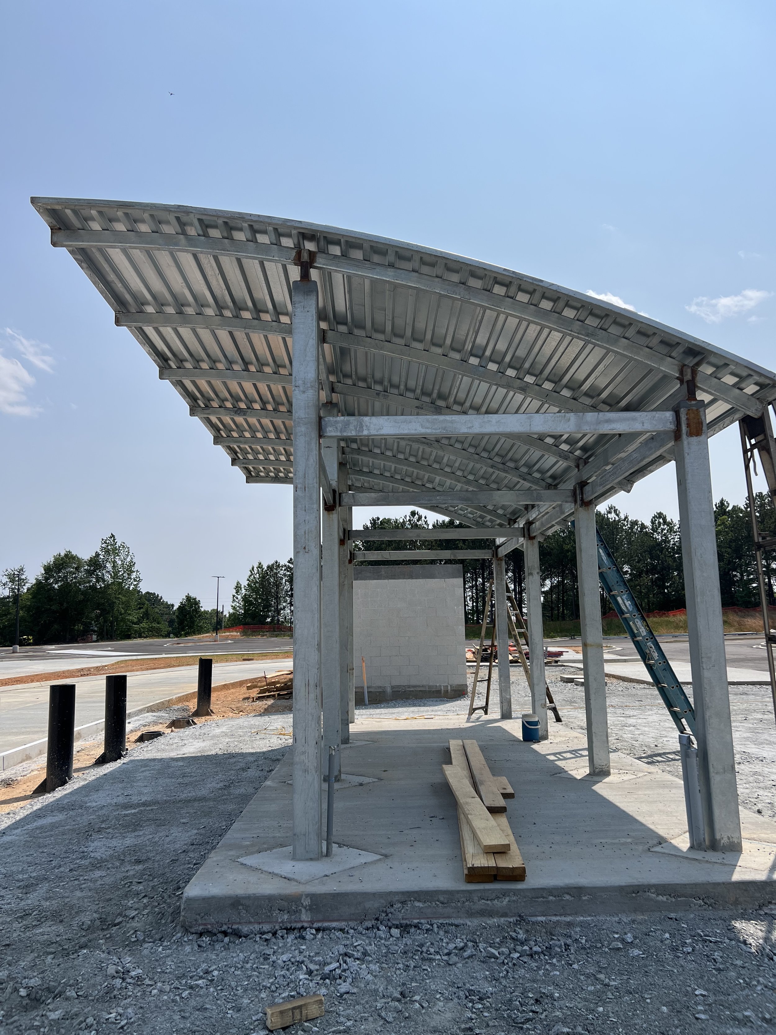 A partially constructed concrete and metal structure with a curved roof, situated on a construction site with scattered lumber and tools. Trees and an open area are visible in the background under a clear sky.