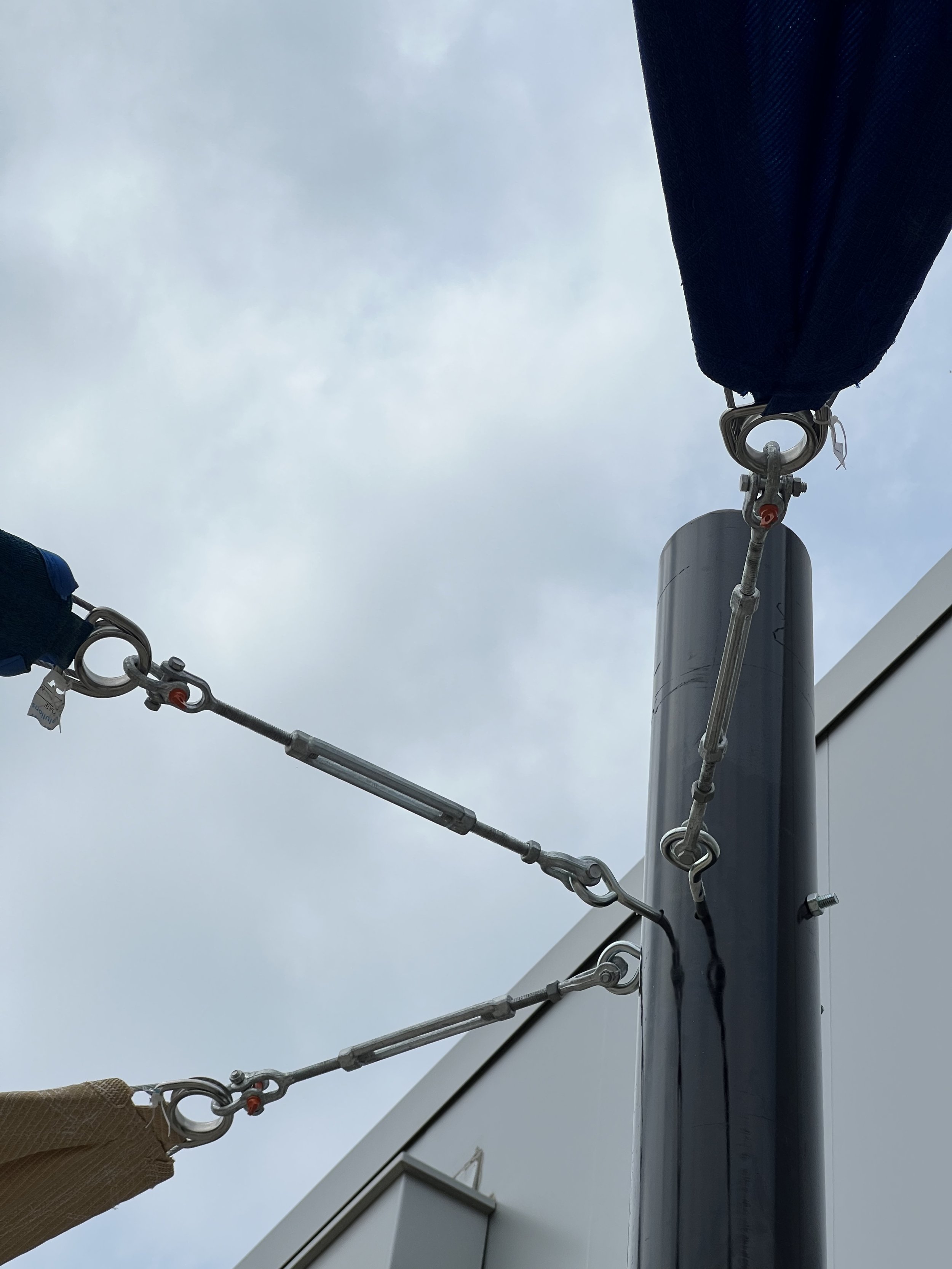 View of tension cables and turnbuckles connected to a metal pole, supporting fabric shades against a cloudy sky.