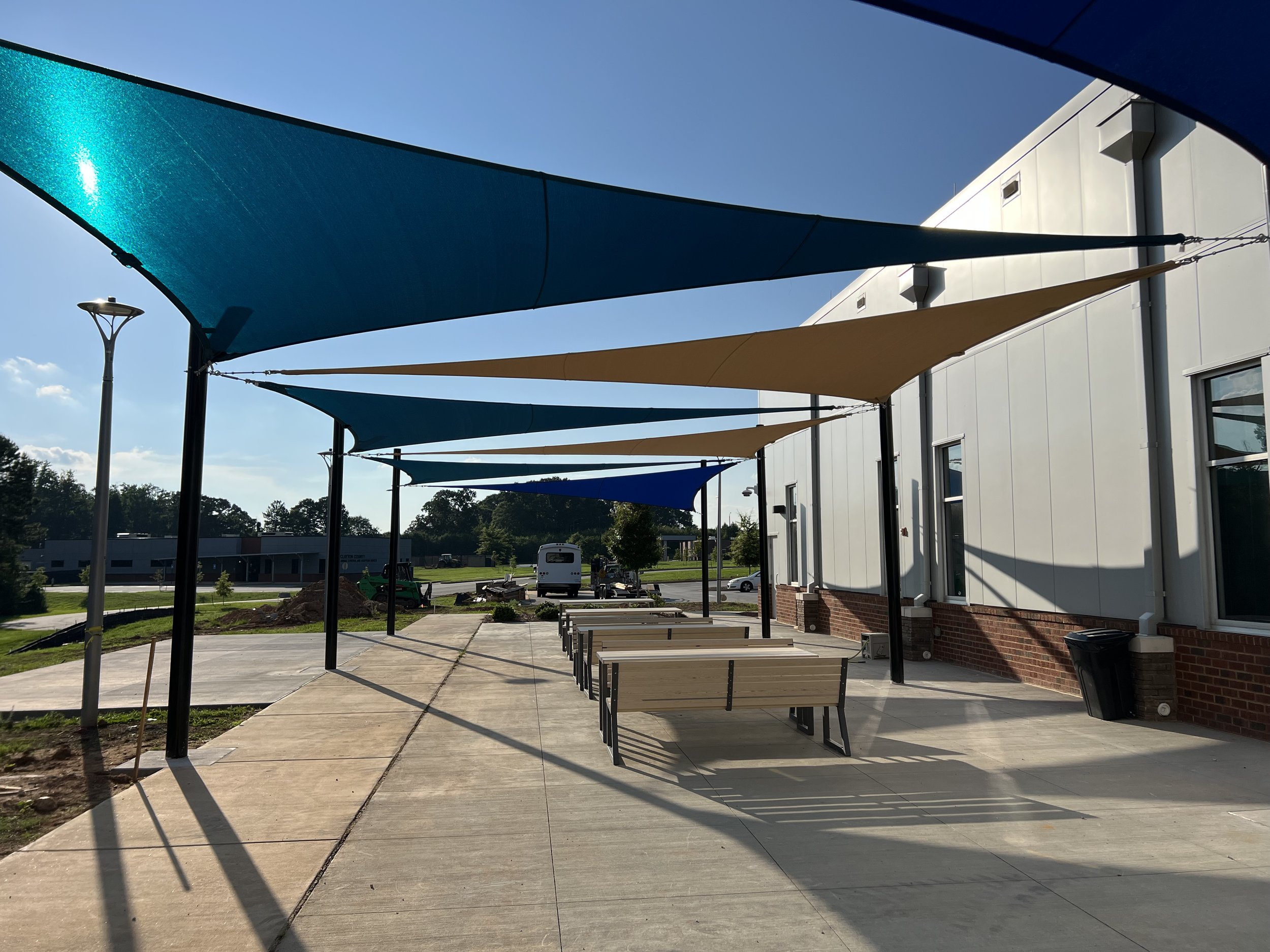 Outdoor seating area with blue and beige shade sails covering benches, adjacent to a building, on a sunny day.