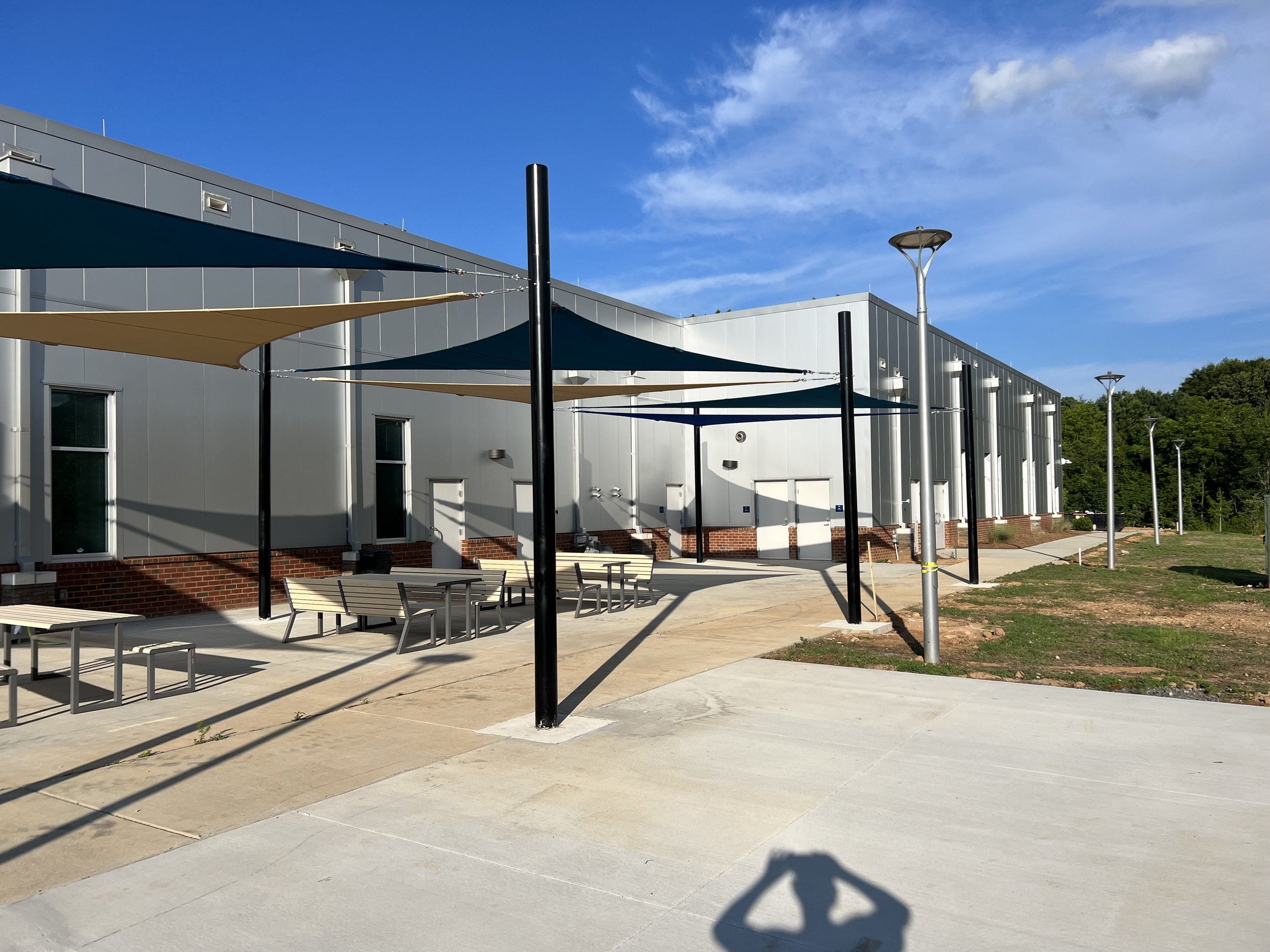 Outdoor seating area with shade canopies and tables in front of a modern building on a sunny day.