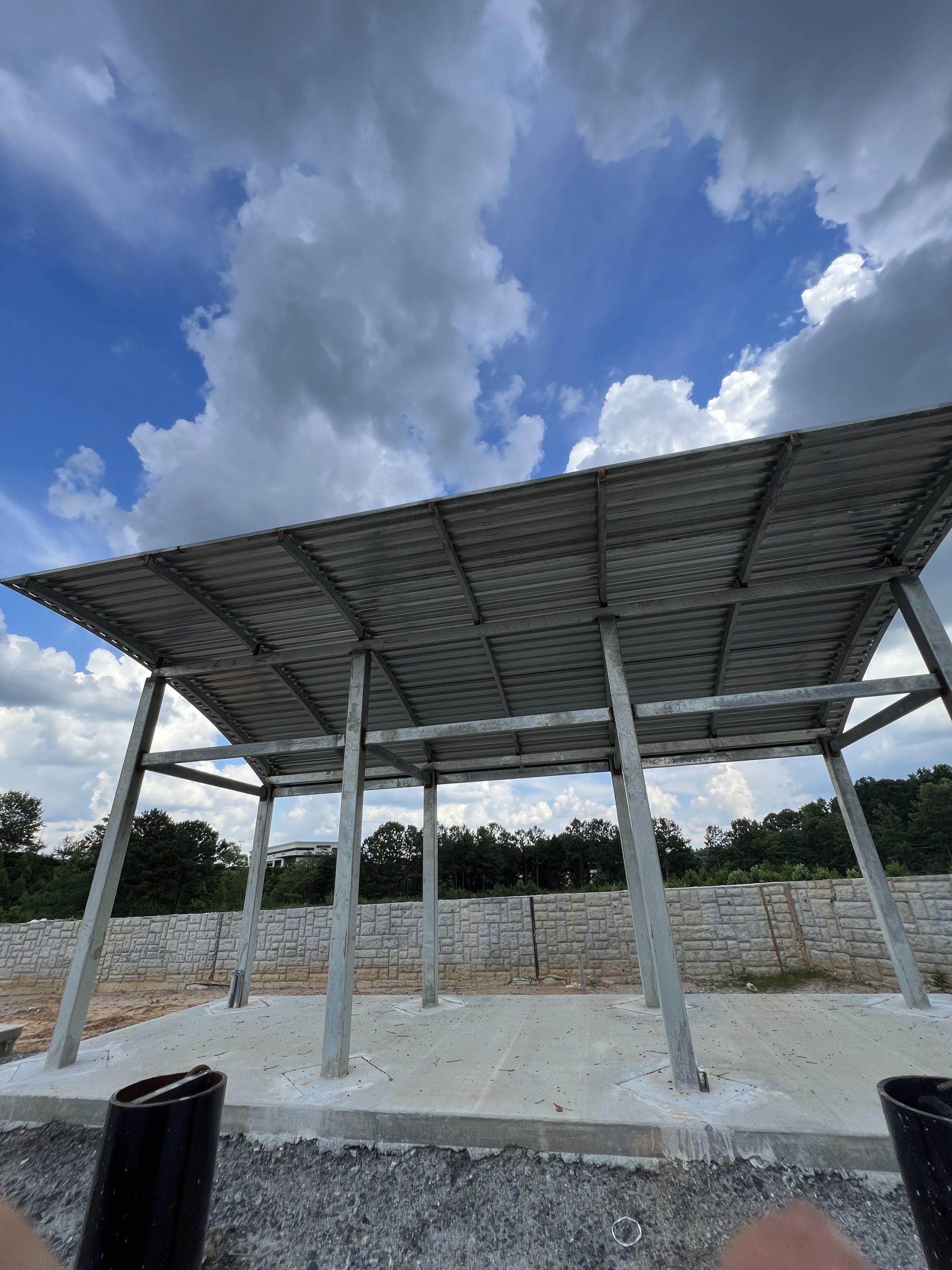 Partially constructed metal canopy with pillars on a concrete slab, against a backdrop of cloudy sky and trees.