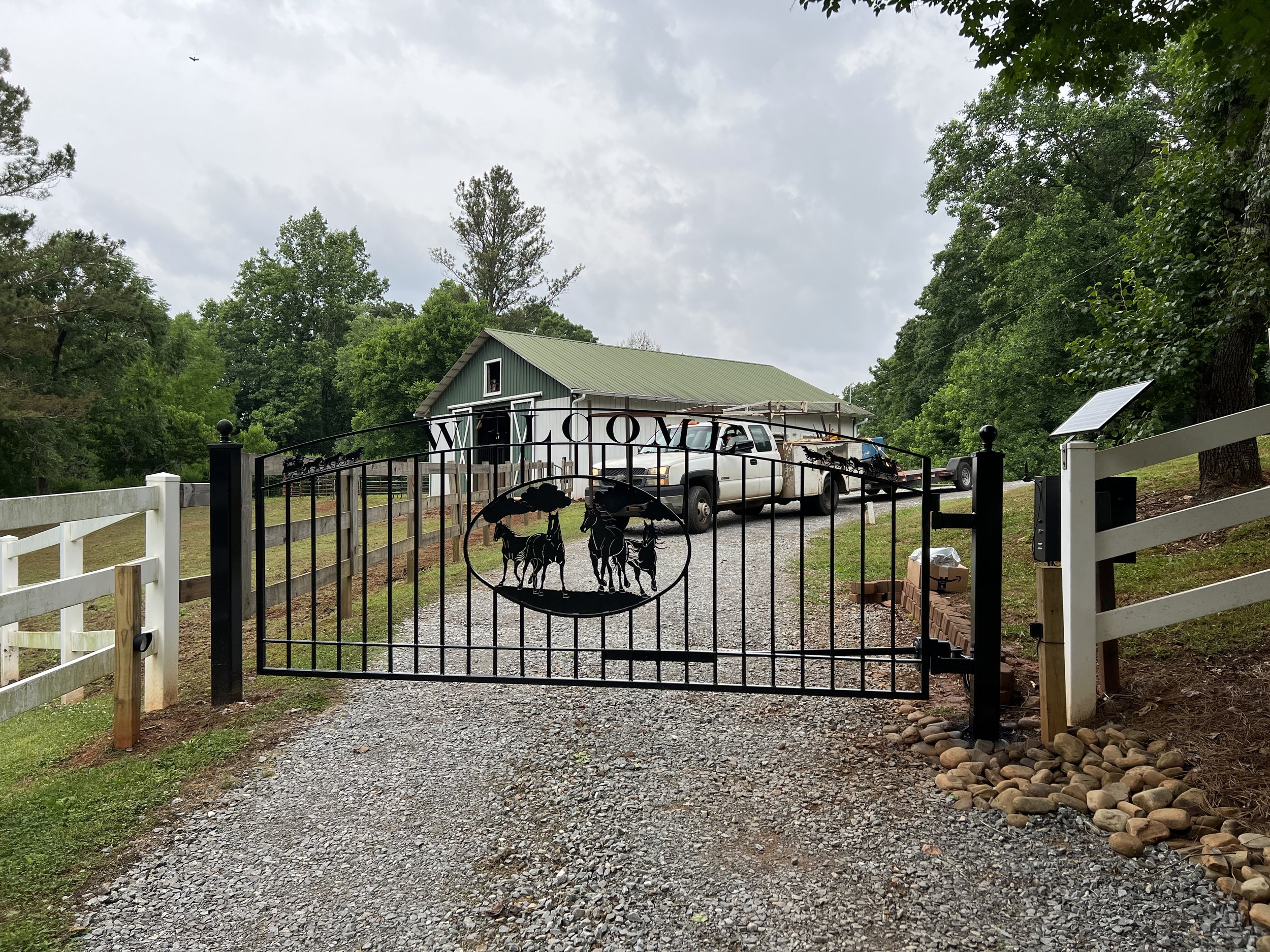 Farm entrance with decorative black metal gate featuring horse design, gravel driveway, green-roofed barn in the background, surrounded by trees.